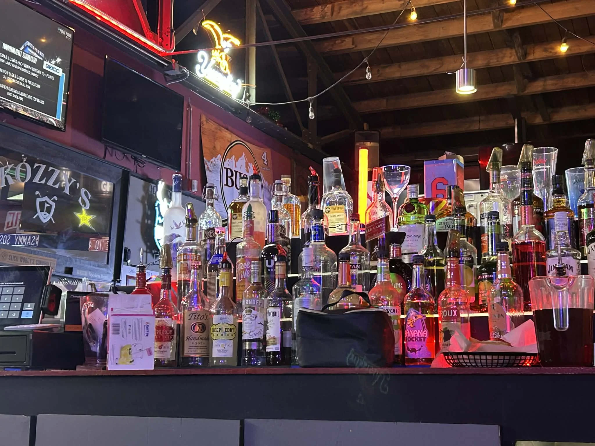 Bar with various bottles of alcohol on the counter, some glasses and bar equipment. Neon signs and a TV screen are also visible in a dimly lit setting.