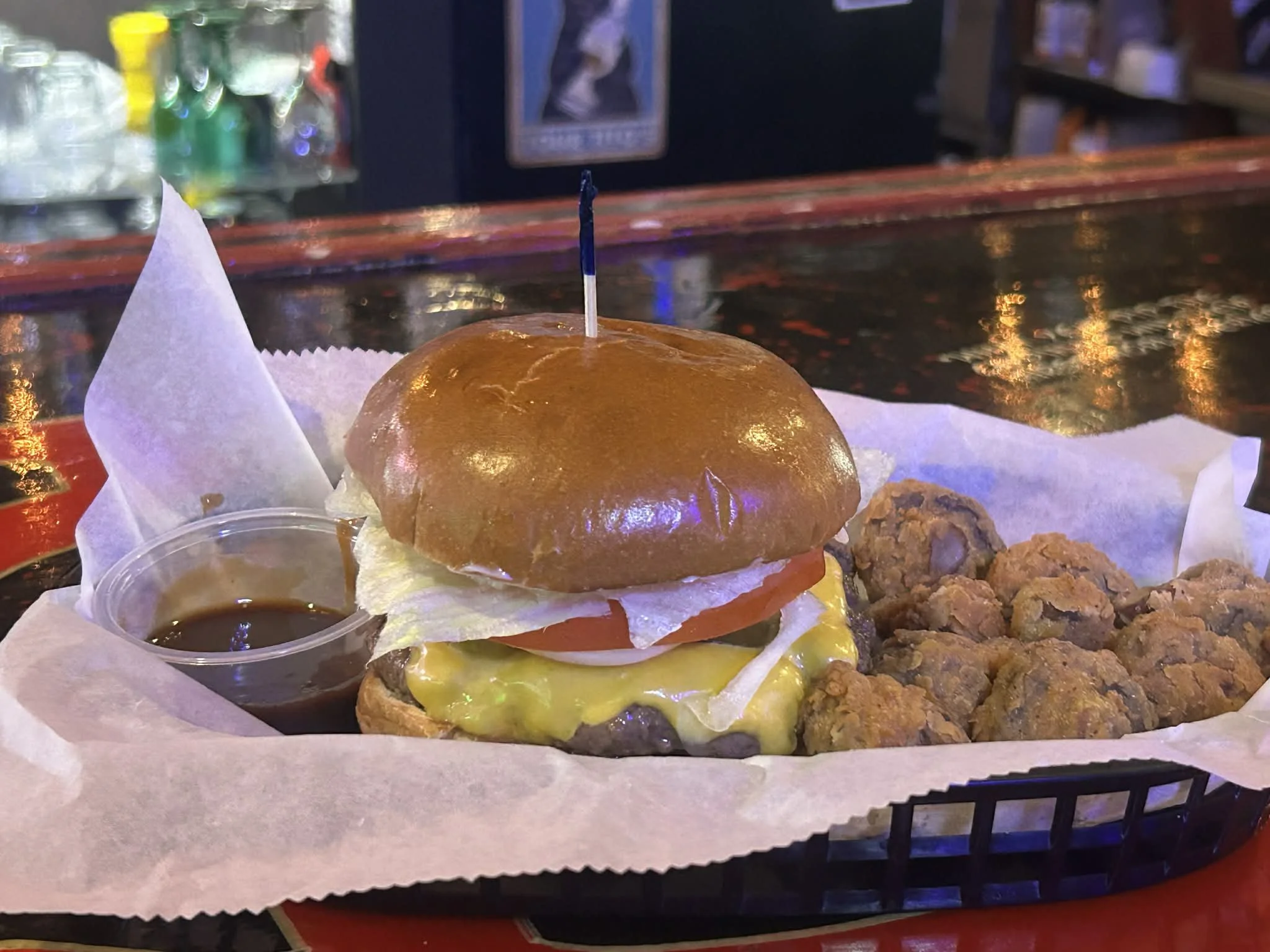 A cheeseburger with lettuce, tomato, cheese, and a bun topped with a toothpick, served with a side of fried bites and a small cup of sauce on a tray lined with parchment paper.