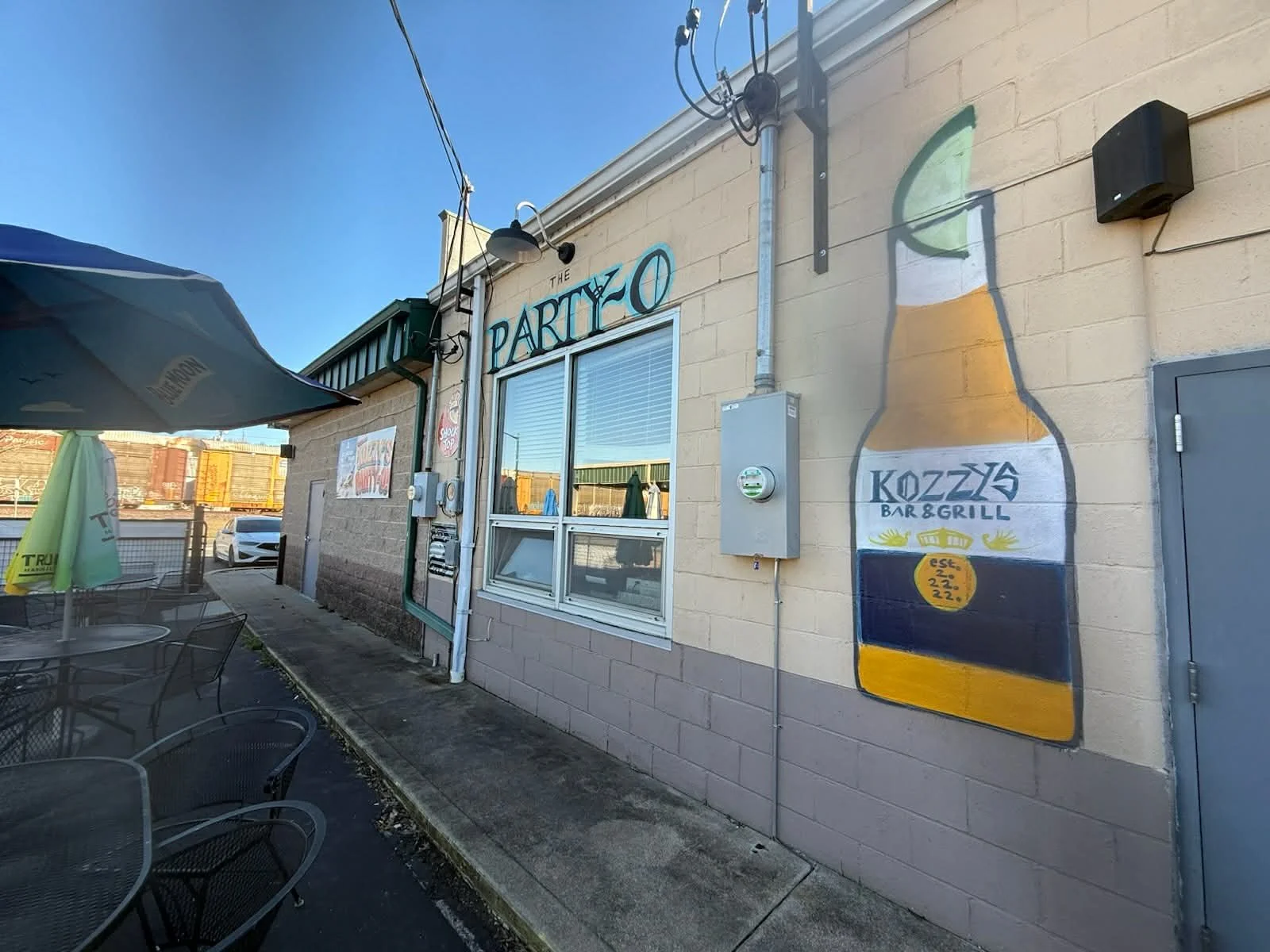 Exterior of a bar and grill called The Party Kozzas, with painted signage of a large beer bottle on the wall and outdoor seating with tables and umbrellas.