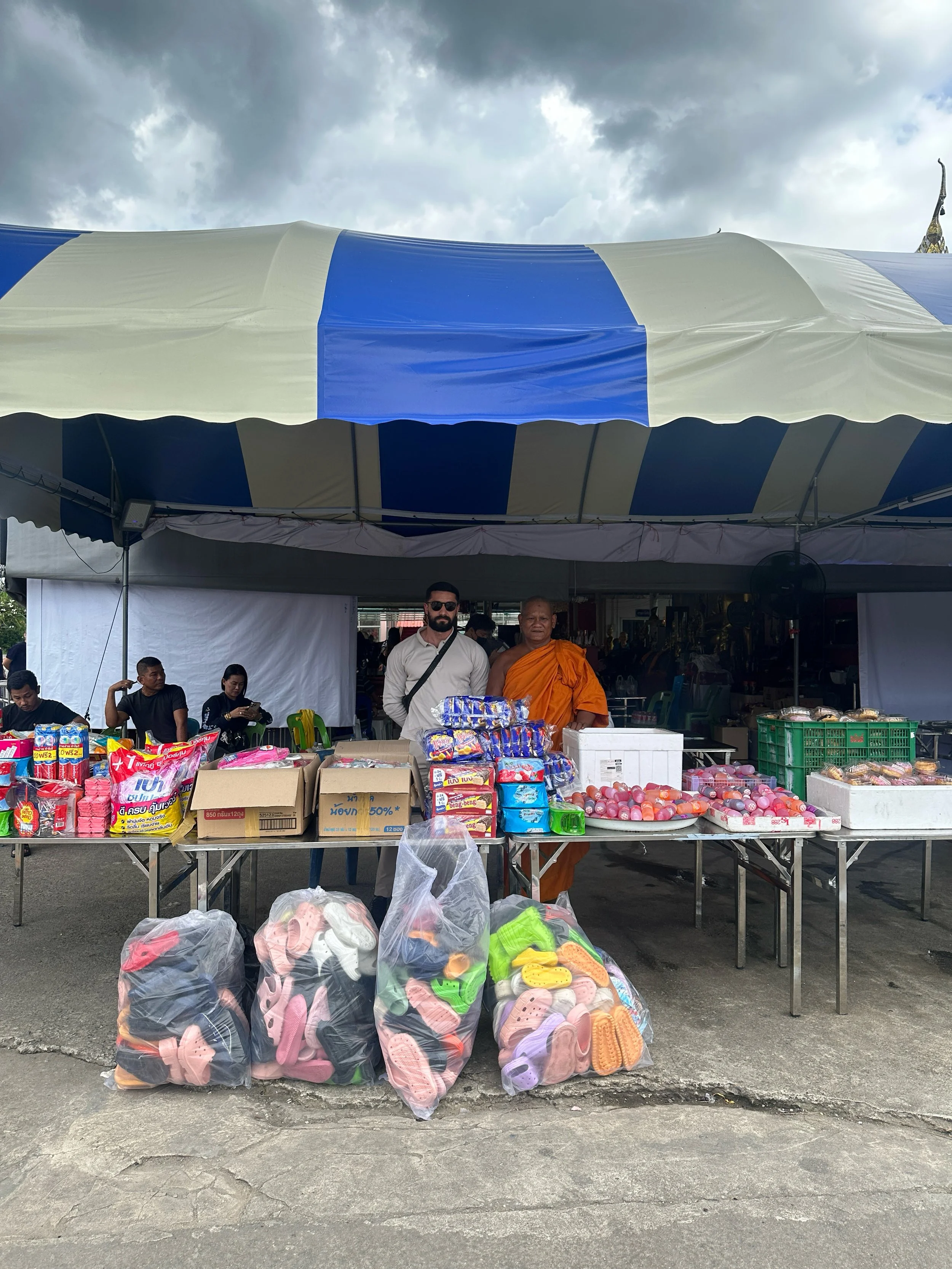 Straßenszene eines Marktes mit einem Verkaufsstand unter einem blauen und weißen Zelt. Es werden Schuhe, Snacks und Süßigkeiten verkauft. Zwei Männer, einer in einer traditionellen orangenen Robe und der andere in weiß, stehen hinter dem Stand. Mehrere Menschen sitzen im Hintergrund.