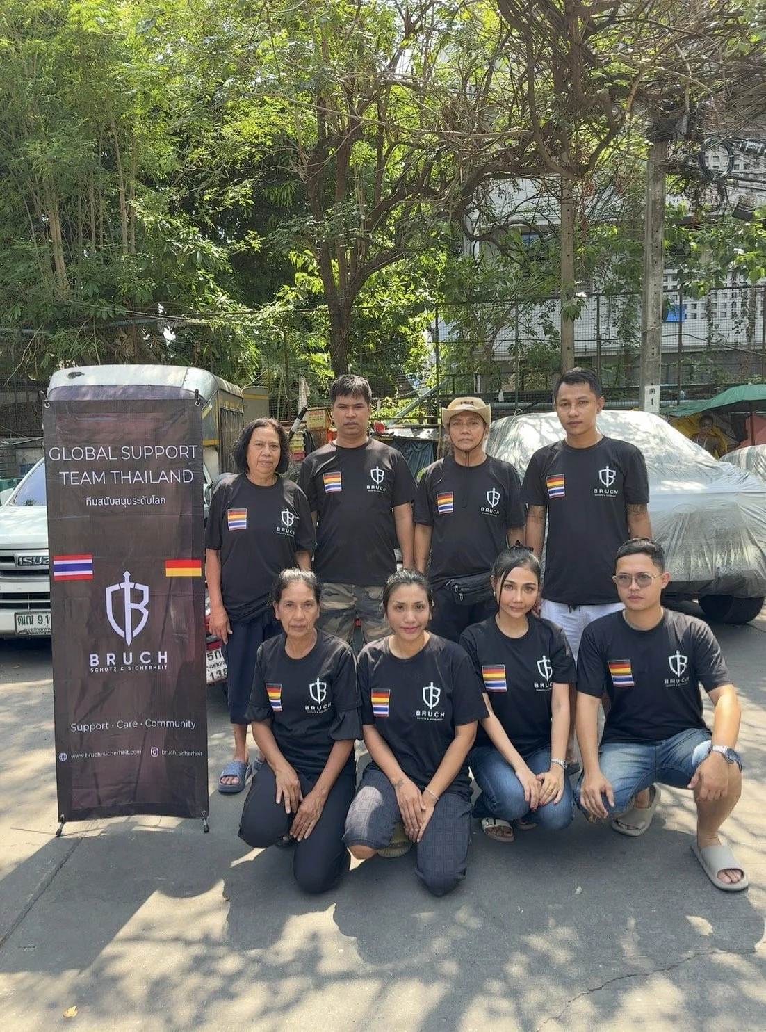 Gruppe von Menschen in schwarzen T-Shirts mit Bruch-Logo, die für den Support in Thailand stehen, vor einem Baum und Fahrzeug, bei Tageslicht.