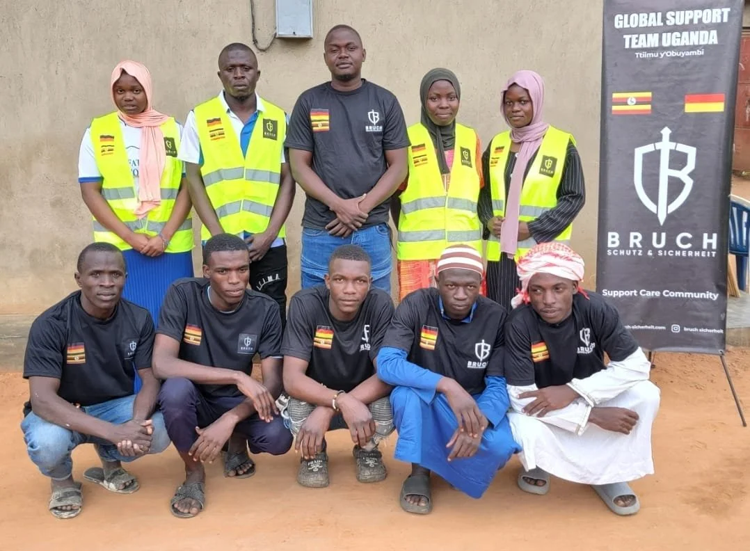 Gruppe von Menschen, einige in schwarzen T-Shirts mit Bruch-Logo und deutschen Flaggen, andere in gelben Sicherheitswesten, vor einer Wand mit Bruch-Banner, Unterstützungsteam Uganda