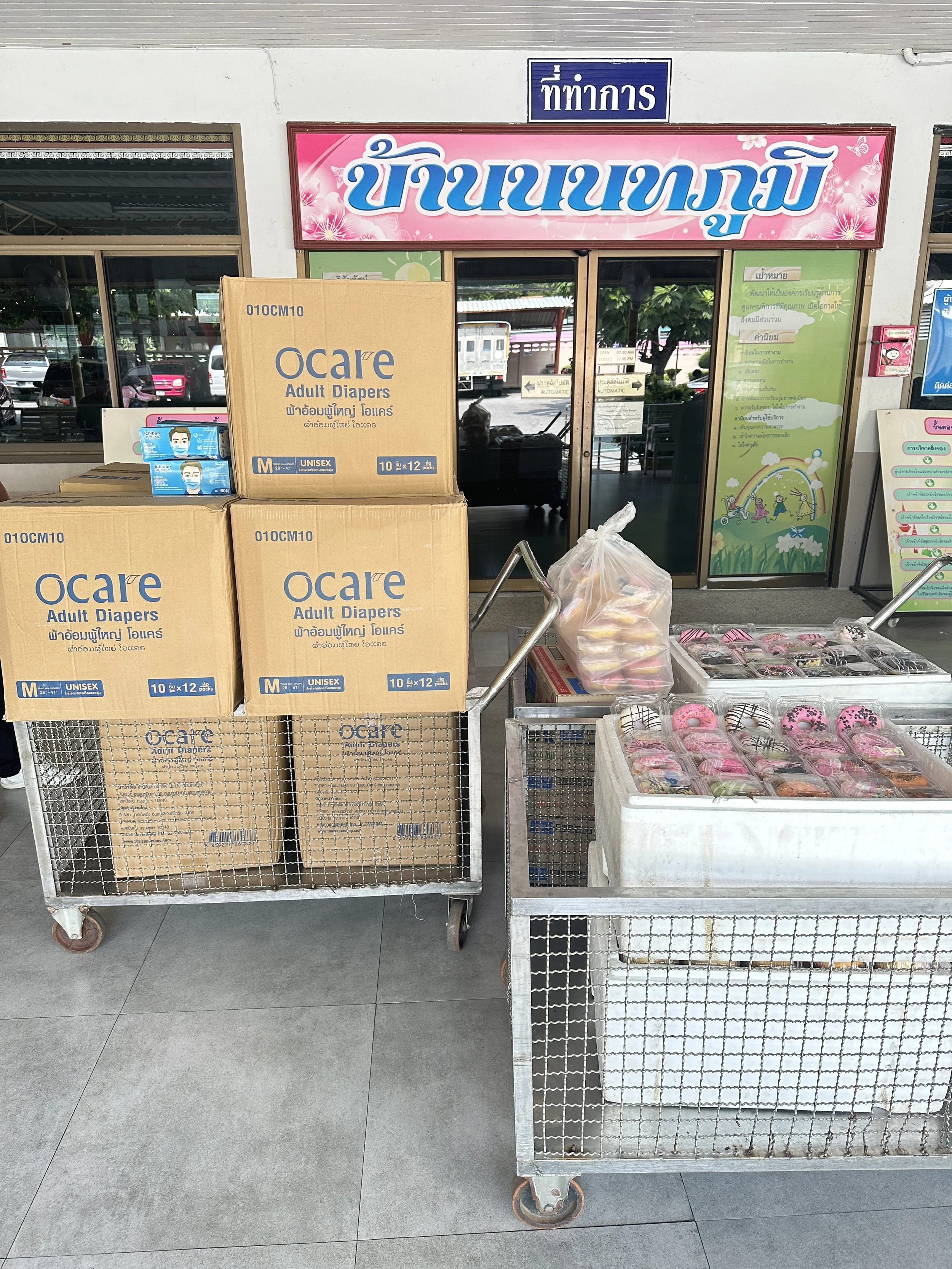 Verkaufstische mit Boxen voller Blumen und Paketen vor einem Ladeneingang mit einem großen pinken Schild und einem blauen Nameschild in einer asiatischen Schrift. Der Hintergrund zeigt einen Eingang mit Glastüren und einige Fahrzeuge.