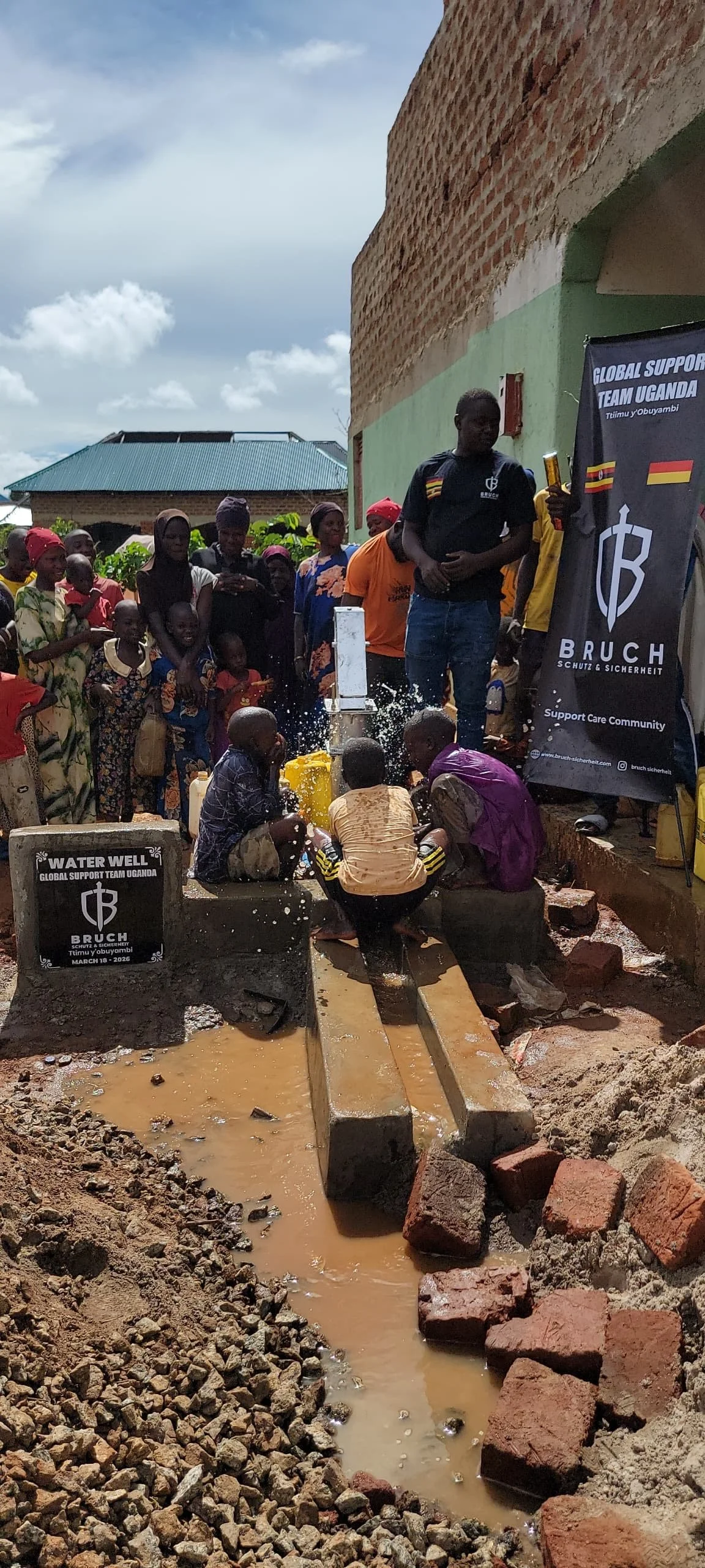 Menschen bei der Wasserbohrung in Uganda, mit Freiwilligen und Gemeinschaft, Wasserquelle, Brach, Sand und Steine, mit Banner von Unterstützungsteam.