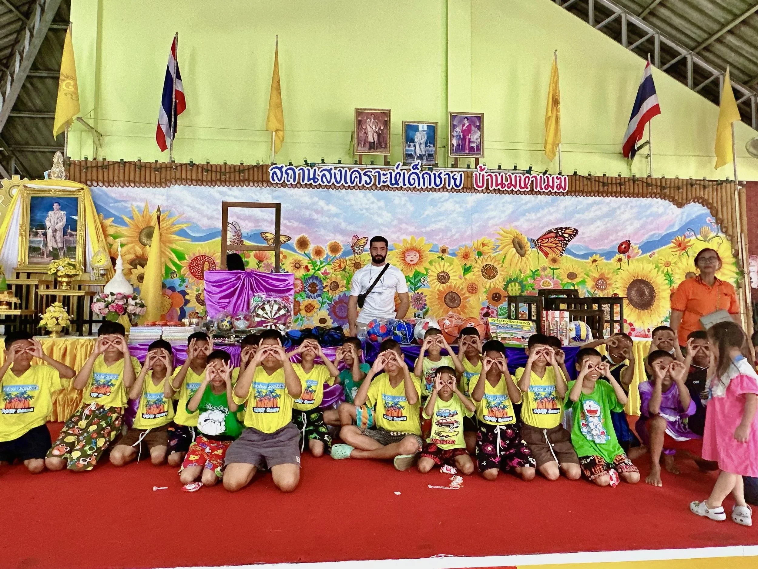 Gruppe von Kindern in bunten T-Shirts, die auf einer Bühne sitzen und die Hände in Form von Ferngläsern vor den Augen positionieren. Im Hintergrund eine bunte Kulisse mit Sonnenblumen, Schmetterlingen und Wolken, sowie ein Portrait von König Bhumibol auf der linken Seite. Zwei Erwachsene stehen auf jeder Seite der Gruppe. Es ist eine festliche Veranstaltung in einem großen Raum.