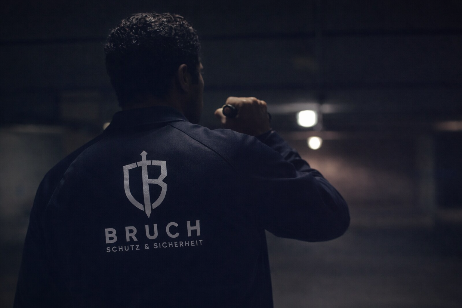 Ein Mann trägt eine dunkle Jacke mit dem Logo und den Schriftzug 'Bruch Schutz & Sicherheit' auf dem Rücken und hält in Hand eine Taschenlampe im dunklen Tunnel.