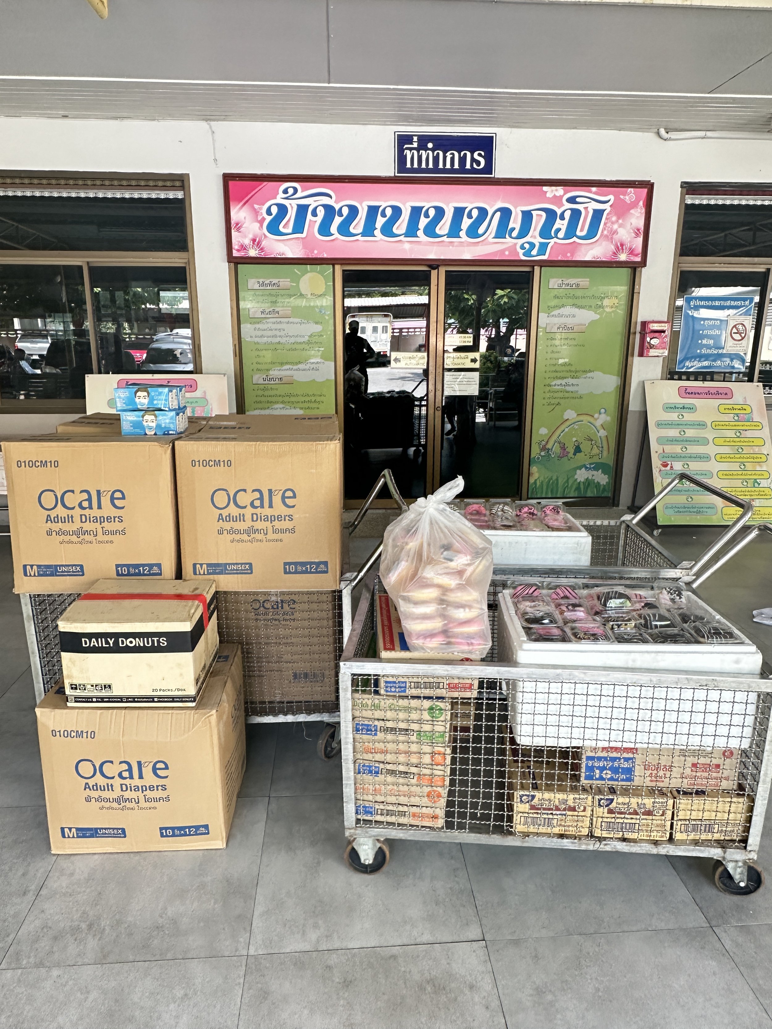 Verkaufstheke mit verschiedenen Produkten vor einem Laden mit Thai-Schriftzug. Im Vordergrund sind Kartons mit Windeln, Donuts und anderen Snacks auf einem Einkaufswagen.