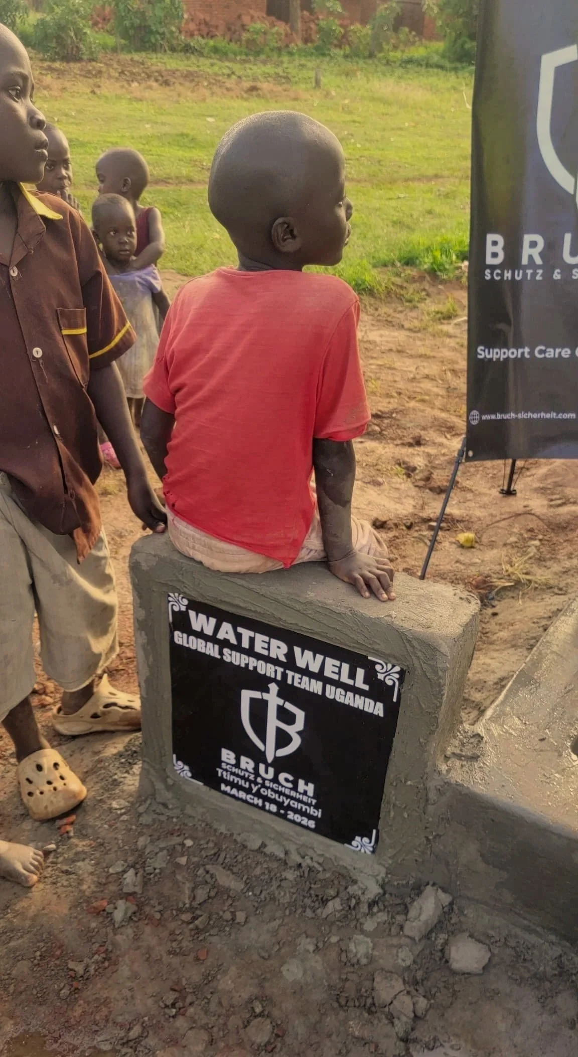 Ein Junge mit dunkler Haut sitzt auf einem Betonblock mit einem Schild, auf dem steht: "WASSERBRUNNEN, Globale Unterstützungsteam Uganda, BRUCH, März 2026." Im Hintergrund sind weitere Kinder und eine grüne Landschaft sichtbar.