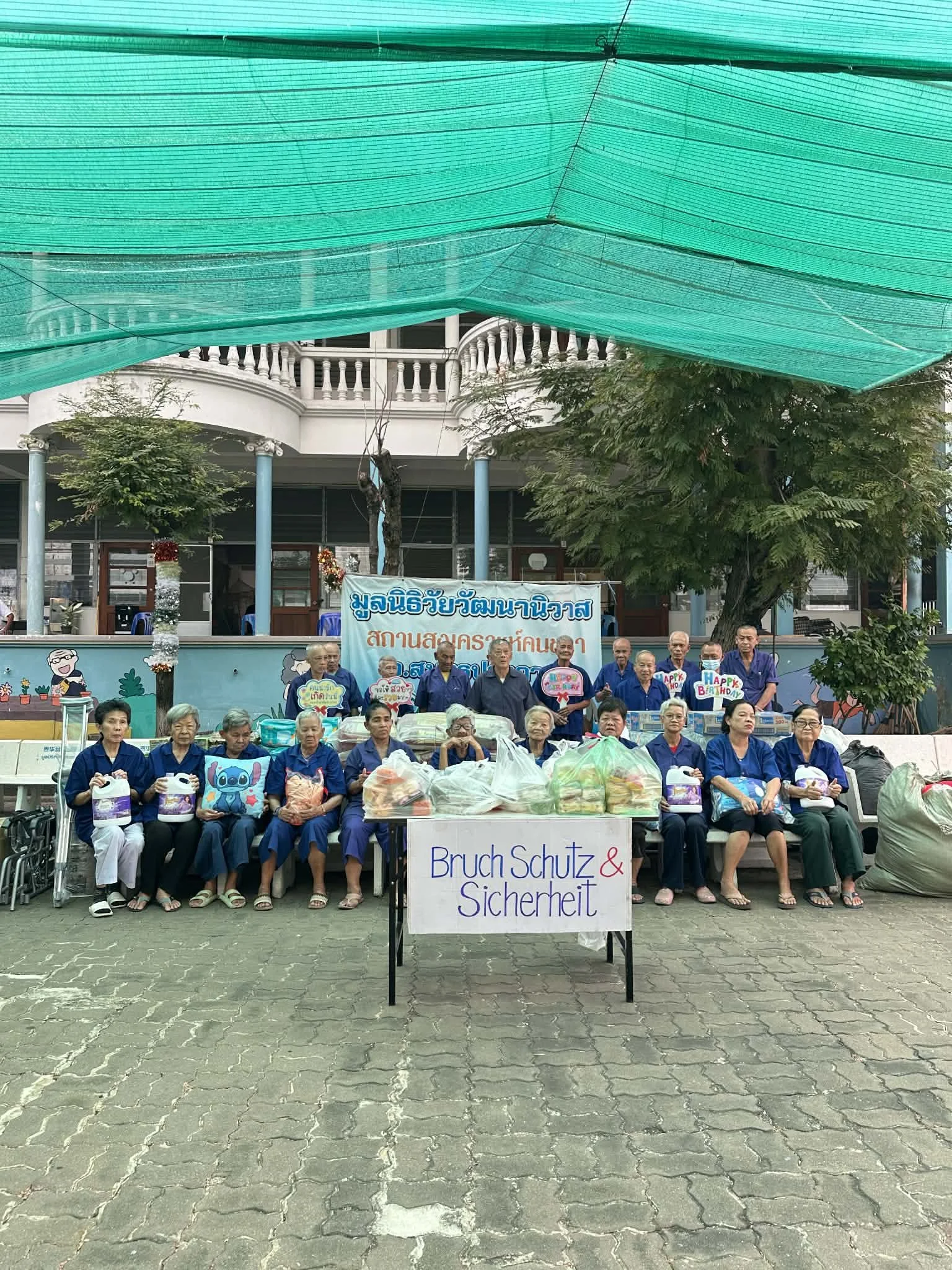 Ältere Menschen bei einem Gemeinschaftsprojekt, das Bruchschutz und Sicherheit fördert, mit einer Tafel und Spenden, in einer outdoor Einrichtung mit Bäumen und einer Gebäude im Hintergrund