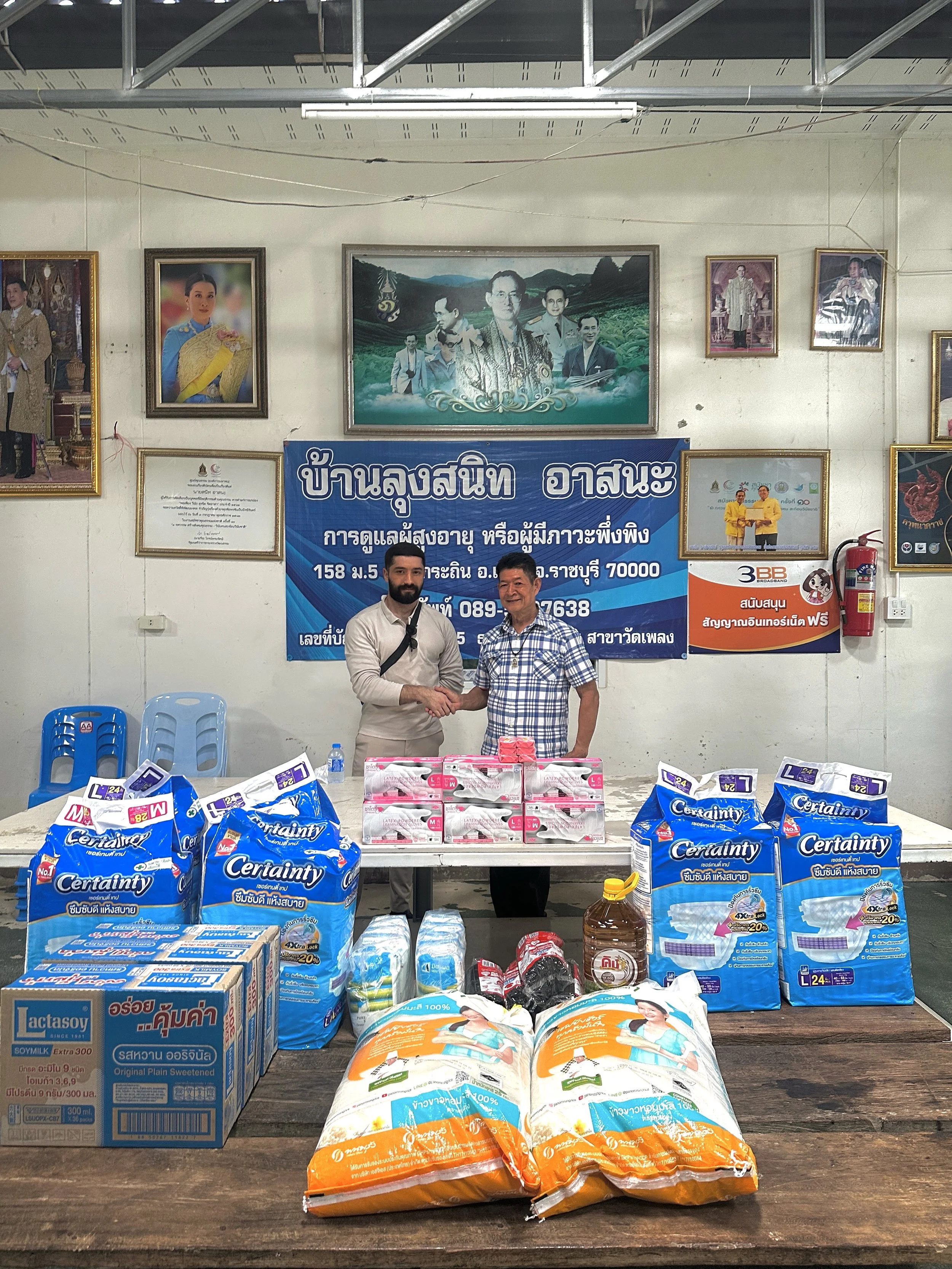 Zwei Männer schütteln sich die Hand hinter einem Tisch mit Lebensmitteln und Haushaltsartikeln. Im Hintergrund ist eine blaue Tafel mit thailändischem Text. Es befinden sich mehrere Bilder an der Wand, darunter ein großes Porträt und kleine Fotos. Es gibt blaue Stühle auf der linken Seite.