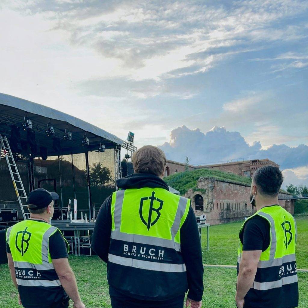 Drei Sicherheitsmitarbeiter in leuchtend gelben Westen stehen vor einer temporären Bühne im Freien, während im Hintergrund eine alte Festung bei Sonnenuntergang zu sehen ist.