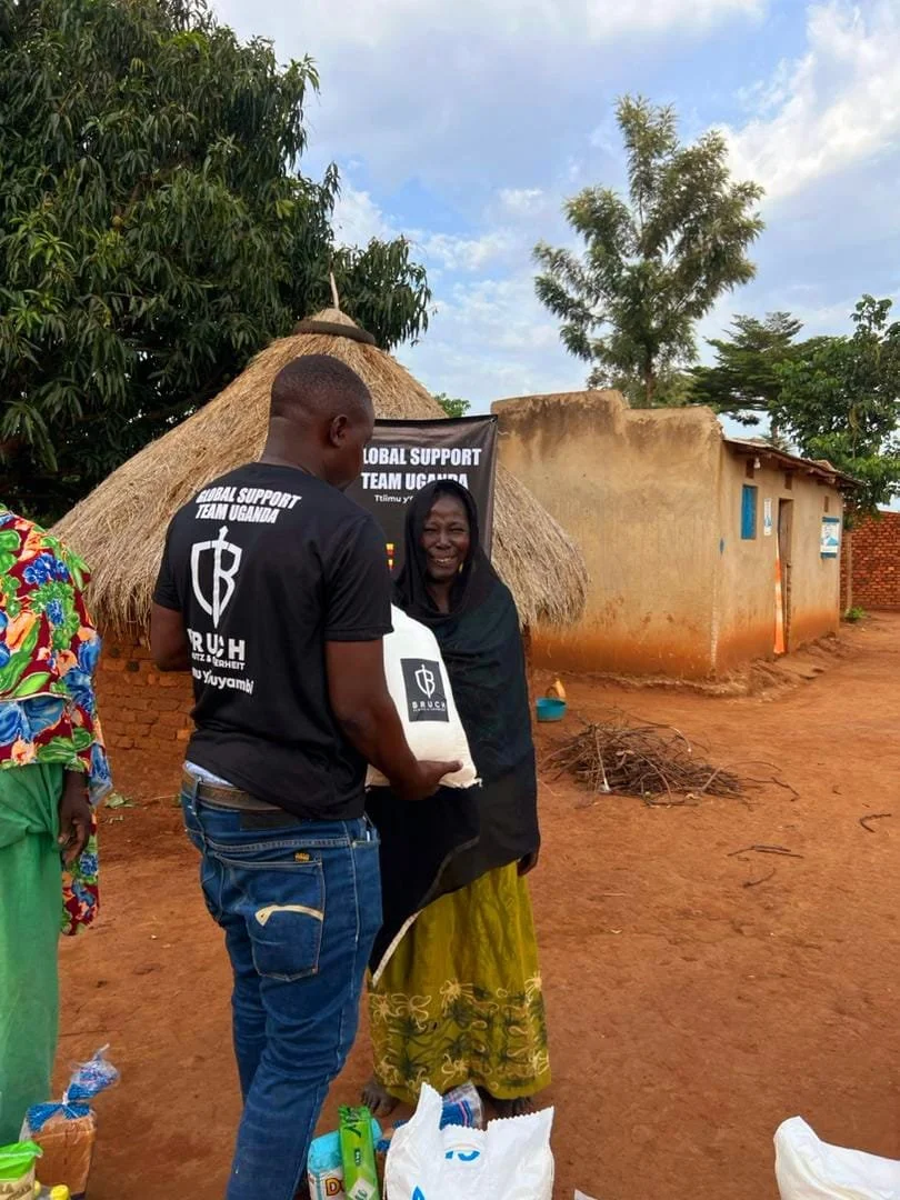 Ein Mann übergibt einer lachenden Frau eine Tüte mit Lebensmitteln oder Hilfsgütern. Sie stehen vor einem traditionellen Hütte in einem ländlichen Gebiet, umgeben von Bäumen und einem sandigen Boden.