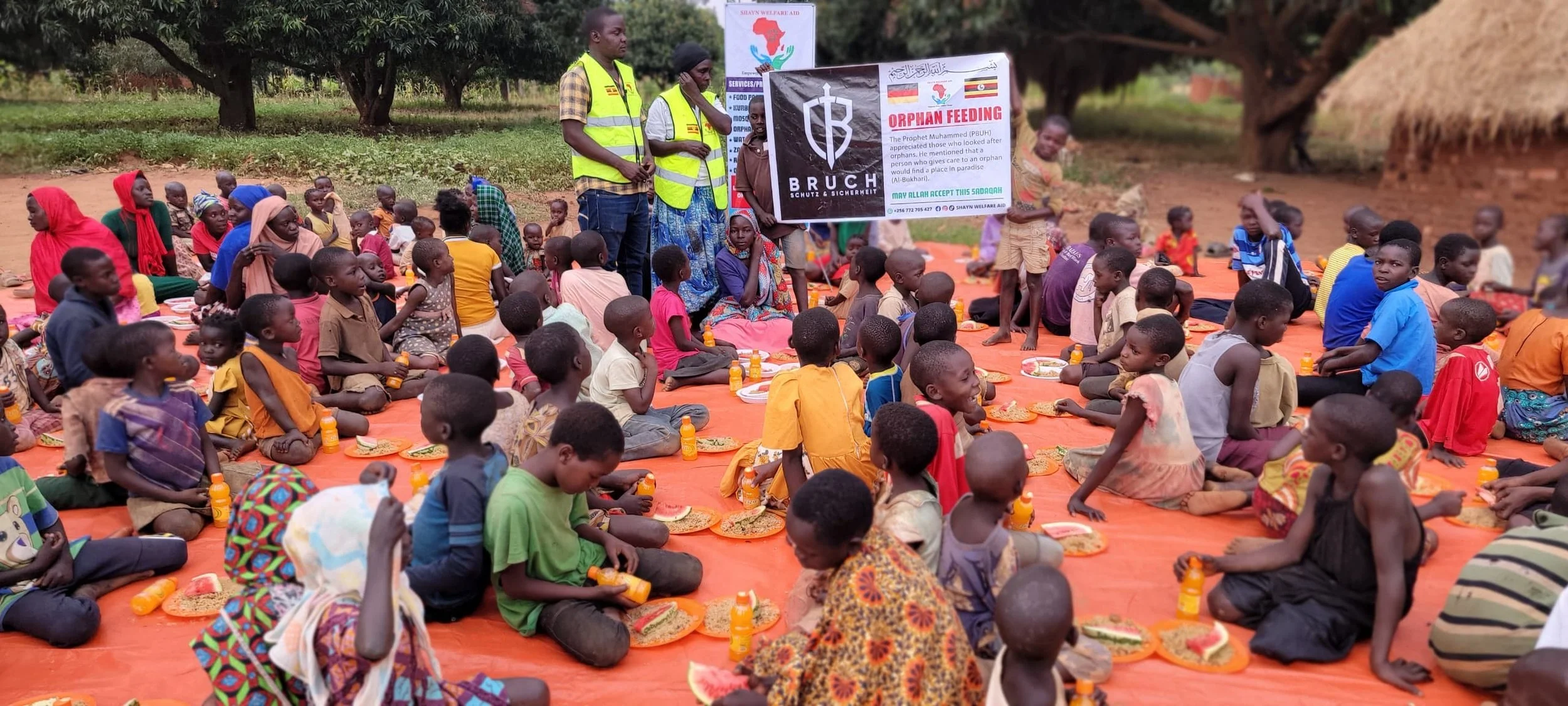 Eine Gruppe von Kindern sitzt auf einer orangefarbenen Matte im Freien und erhält Mahlzeiten. Einige Kinder halten Flaschen, andere haben Teller vor sich. Es gibt Männer, die Plakate halten, eines mit einem schwarzen Symbol und einem anderen mit Flags und Text, die auf eine Waisenhaus- und Versorgungshilfe hinweisen.