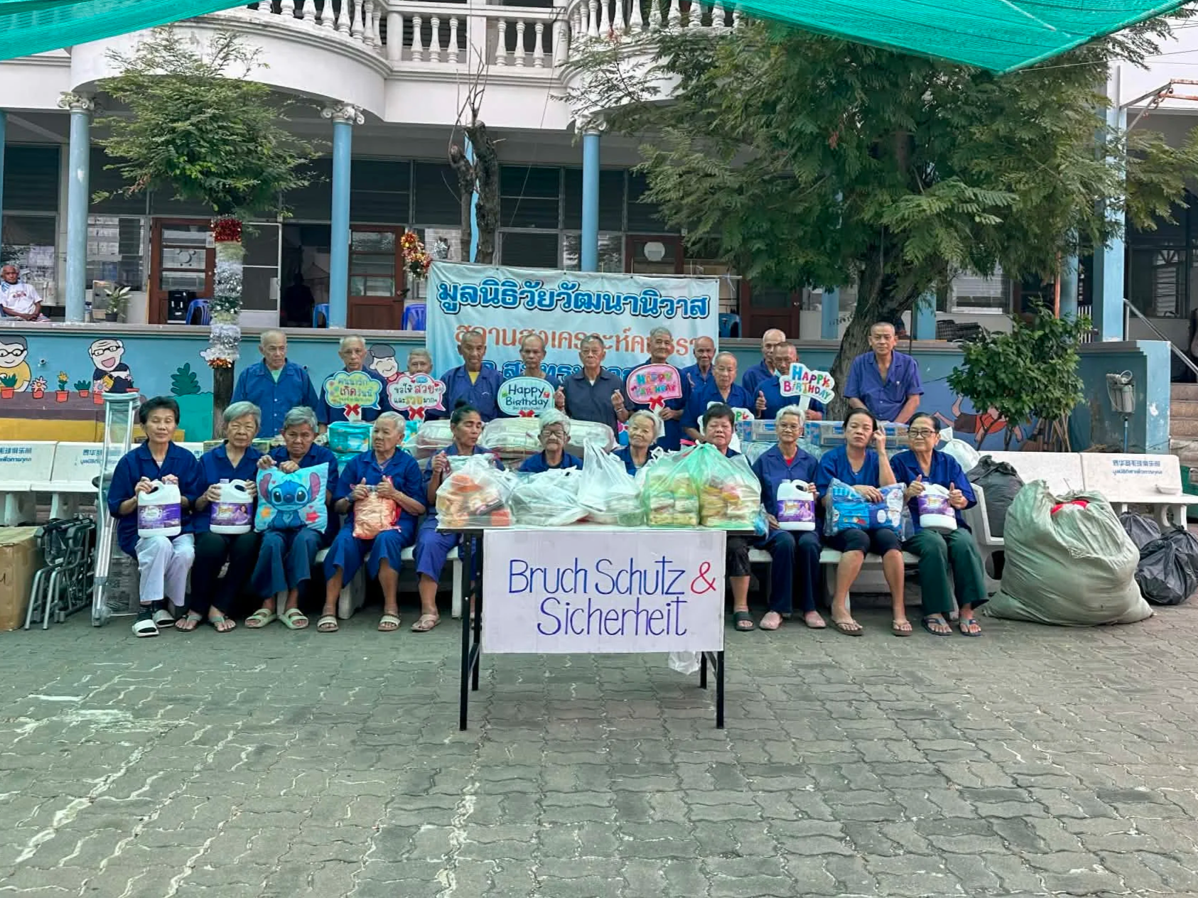 Gruppe älterer Menschen sitzt auf Bänken mit Spenden und Geschenken vor einem Gebäude. Es gibt Schilder mit Glückwünschen und einem Banner mit Text in deutscher Sprache. Die Gruppe trägt blaue Kleidung.