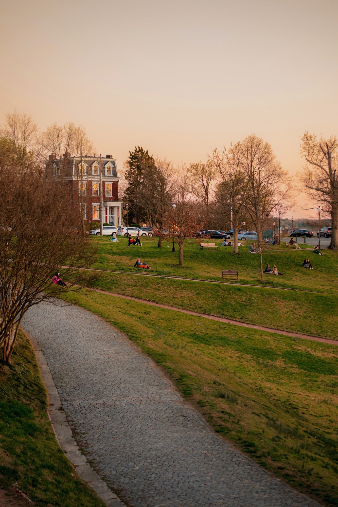 A peaceful park scene during sunset with a winding stone pathway, leafless trees, people sitting and enjoying the scenery, and a large house in the background with cars parked nearby.