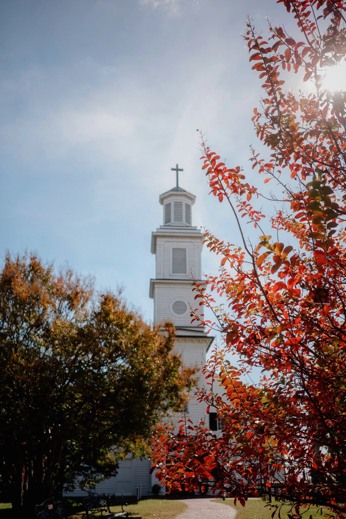 A church steeple with a cross on top is seen behind trees with red and orange leaves, on a sunny autumn day with a clear blue sky.