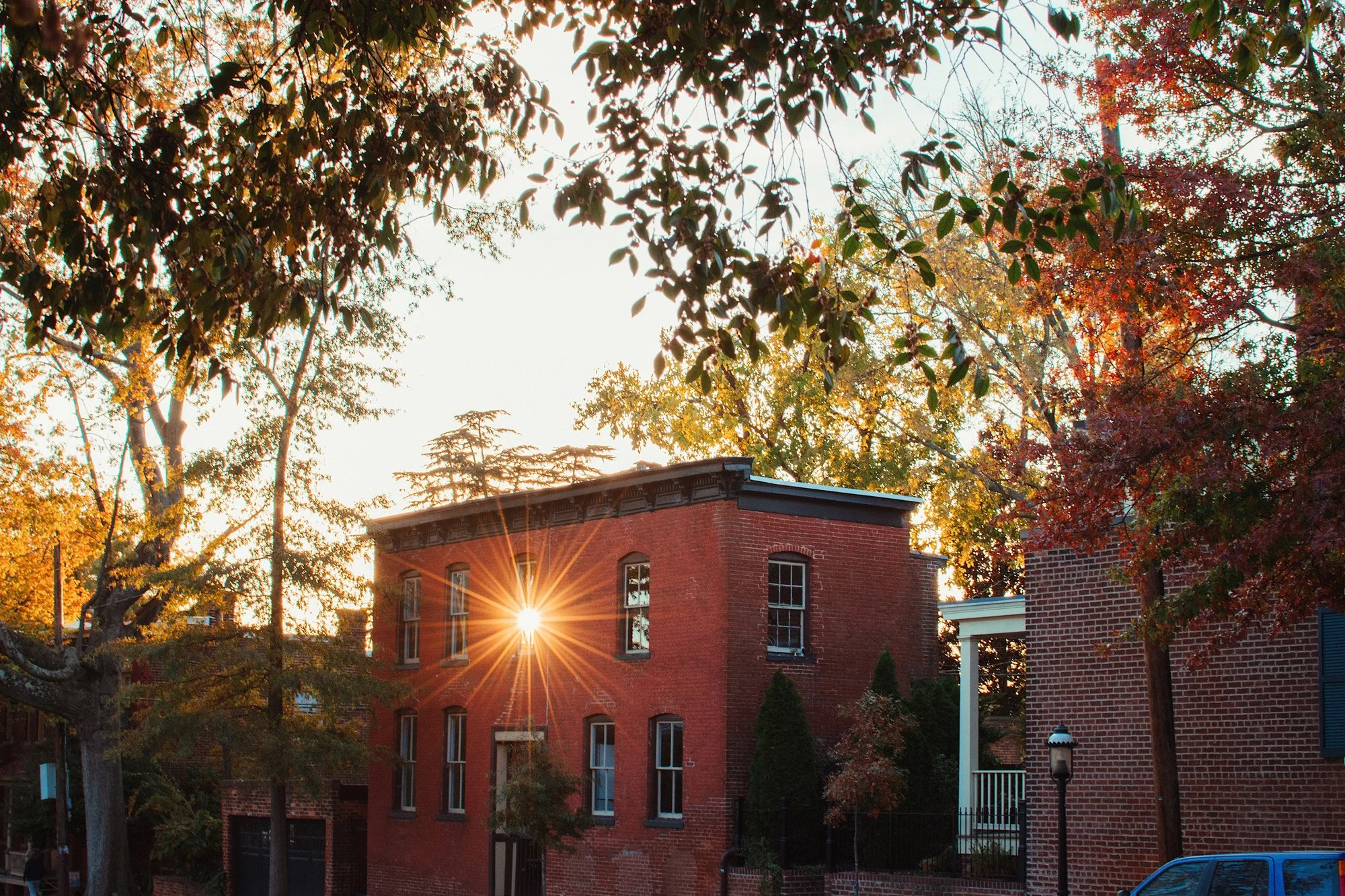 Sunset over a brick building surrounded by trees with fall foliage, sunlight peeking through windows and branches.