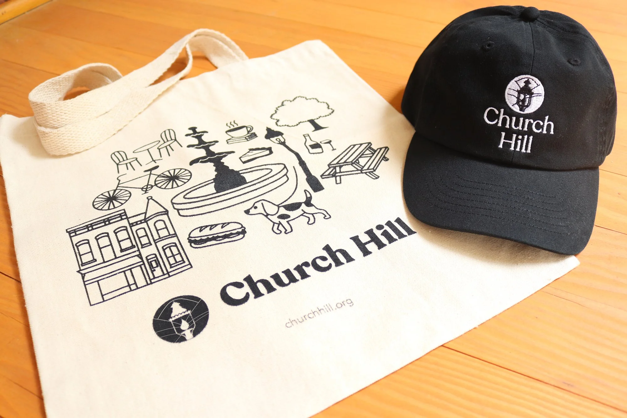 A cream tote bag and a black cap with 'Church Hill' embroidered on them. The tote bag features line art illustrations of a fountain, building, tree, coffee cup, dog, bicycle, picnic table, and a burger, along with the website 'churchhill.org' and a logo of a globe with a cross.