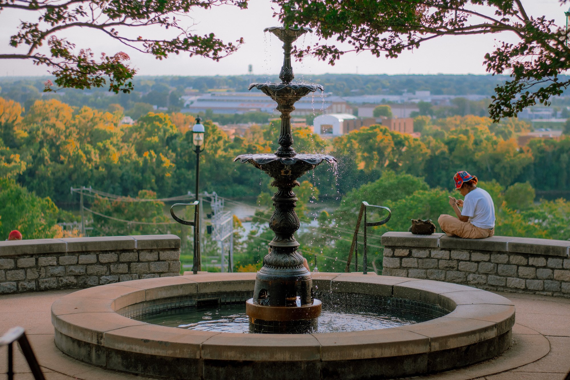 A decorative fountain with multiple tiers in a park, framed by overhanging tree branches, with a person sitting nearby using a phone and greenery extending into the distance.
