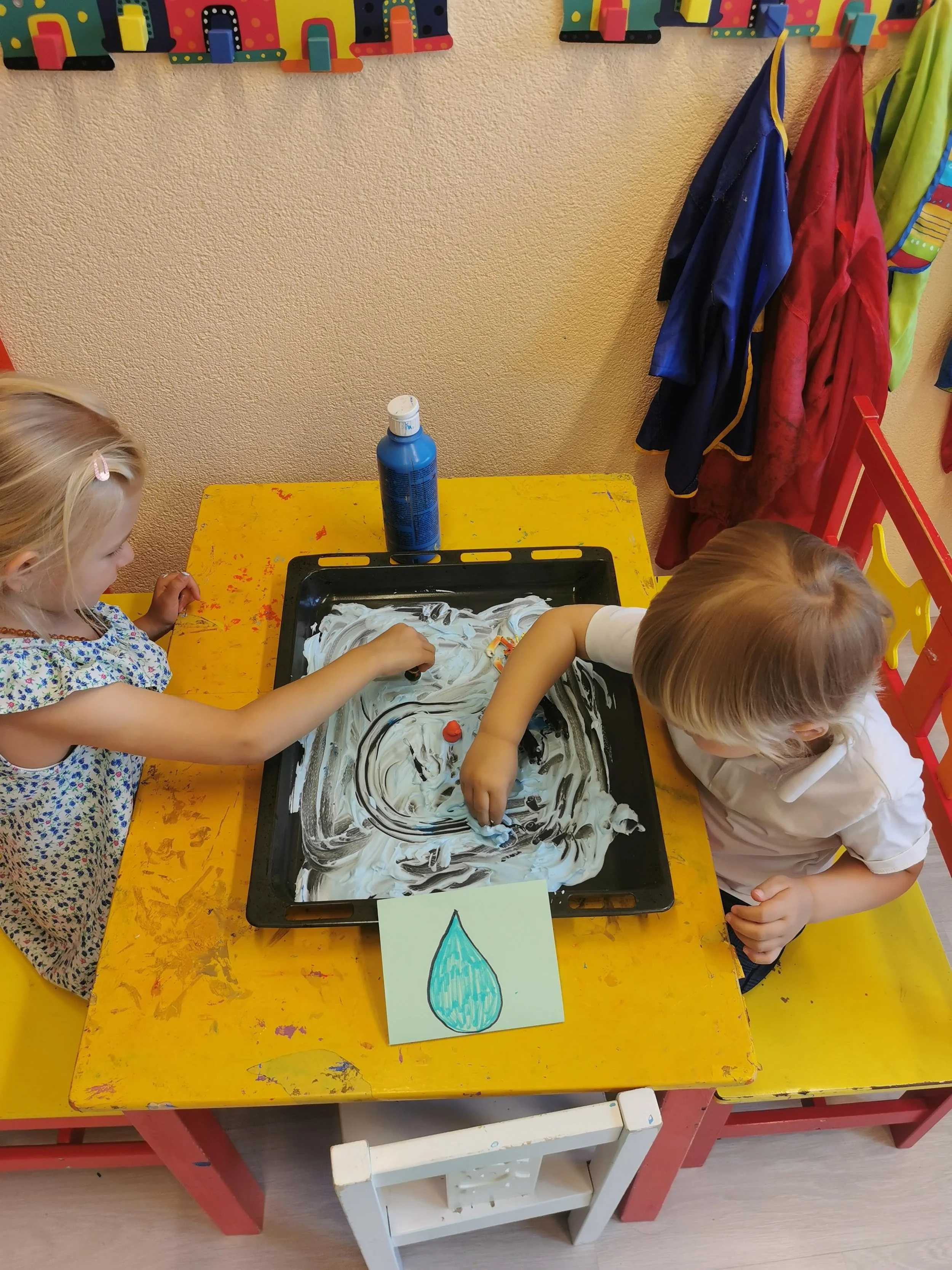 Two young children, a girl and a boy, are engaging in a sensory activity with a tray of swirling soap foam, using colored items. The girl is on the left side of the table, and the boy is on the right. A drawing of a water droplet is taped to the front of the table.