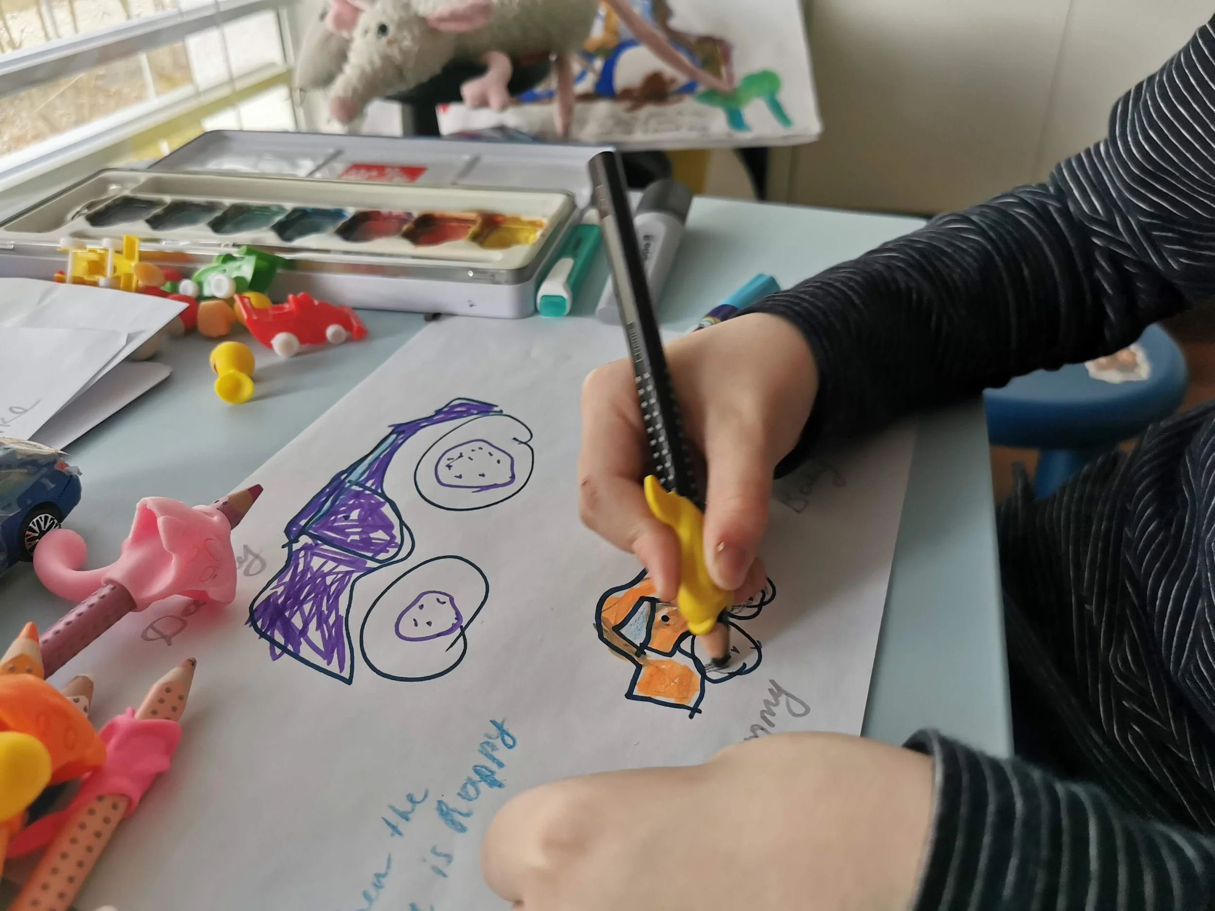 A person drawing colorful illustrations on a white sheet of paper with markers and toys scattered around a desk.