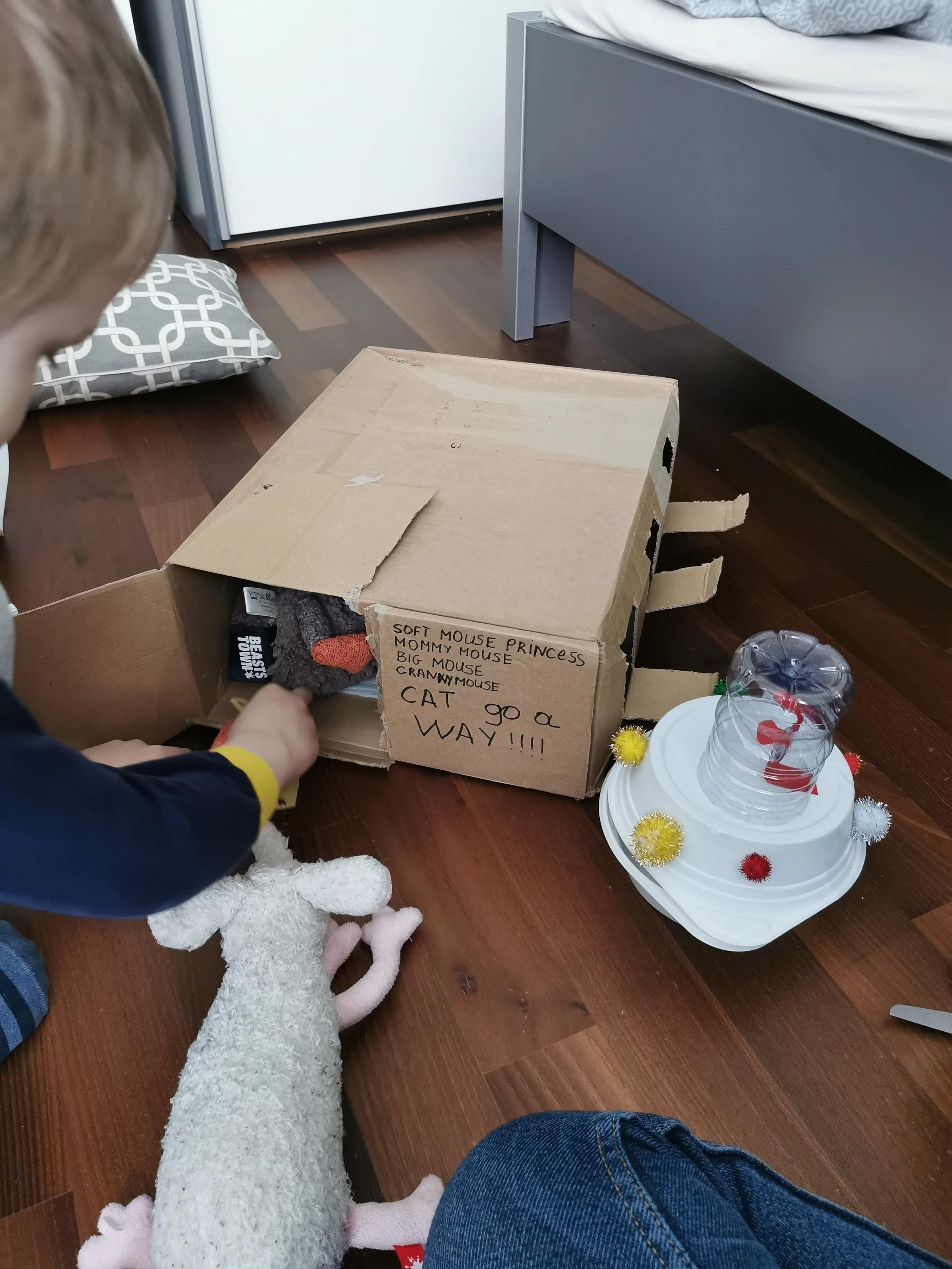 A child placing a stuffed mouse toy into a cardboard box labeled with different mouse names and a note about a cat. The box is on a wooden floor, next to a stack of white plastic plates with a clear plastic cup on top. There is a gray bed with a pillow and a gray drawer in the background.