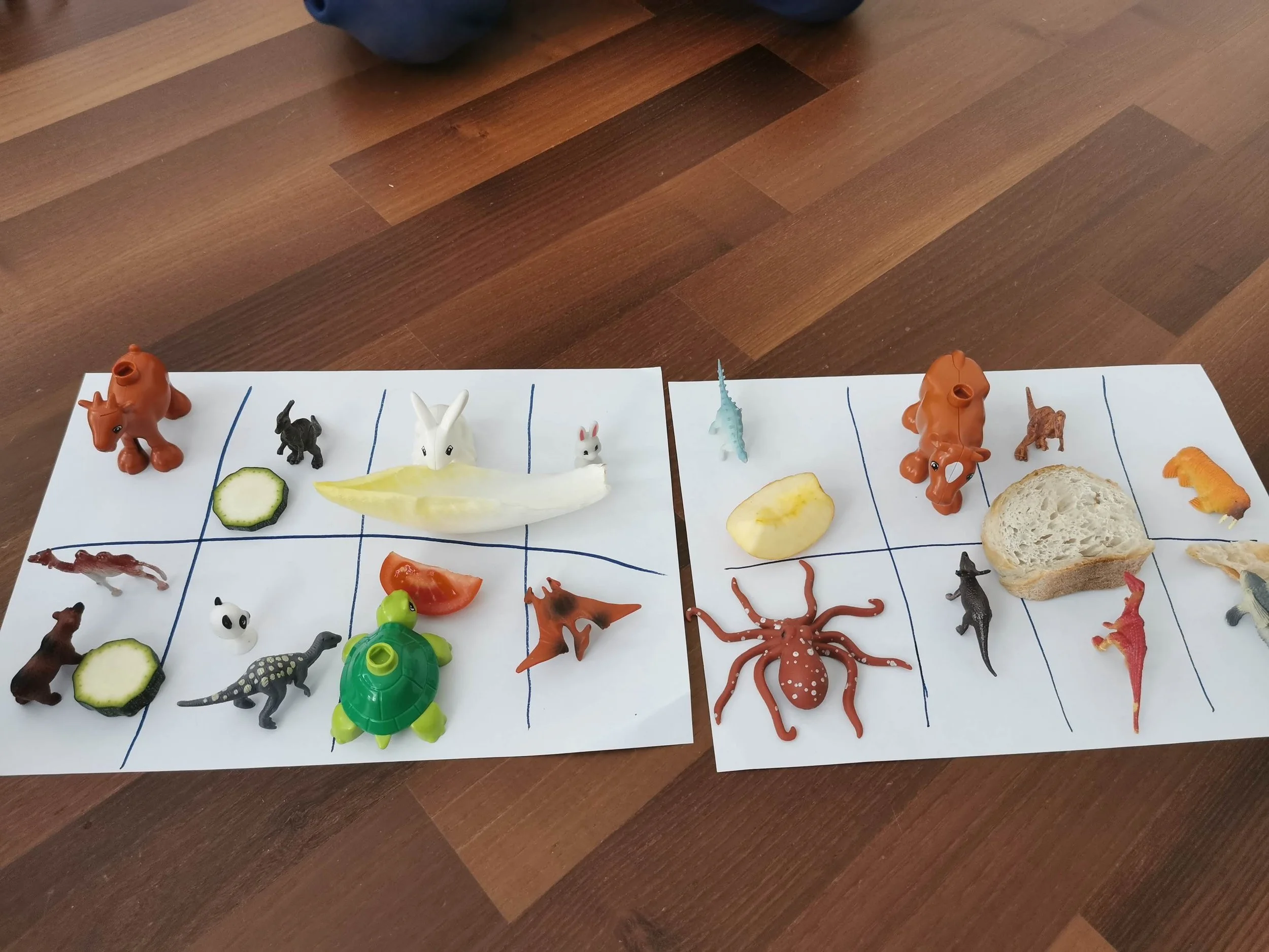 Two sheets of white paper with blue lines and various toy animals, vegetables, and food items arranged on them, on a wooden floor.