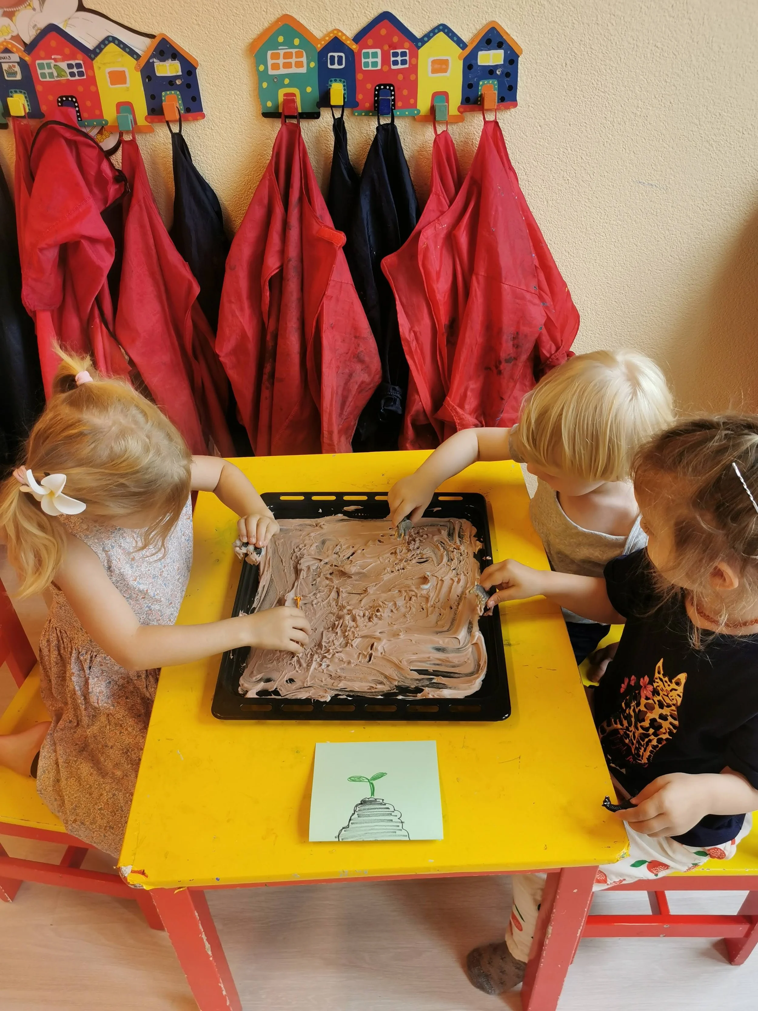 Three children painting on a tray of brown paint at a yellow table indoors, with colorful house-shaped hooks and red smock coats hanging on the wall behind them.