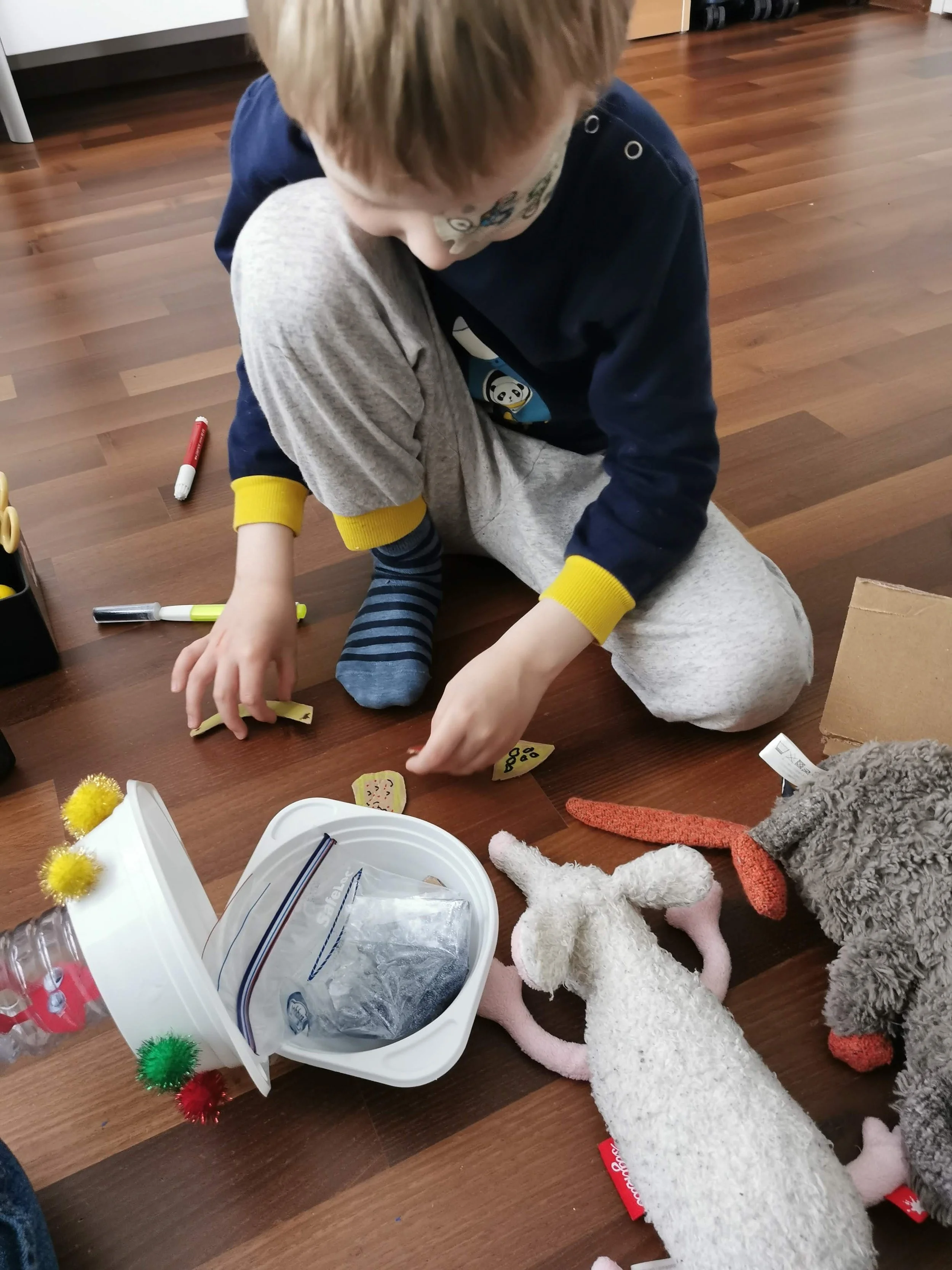 A young boy playing with colorful fabric pieces and stuffed animals on a wooden floor, surrounded by markers and craft supplies.