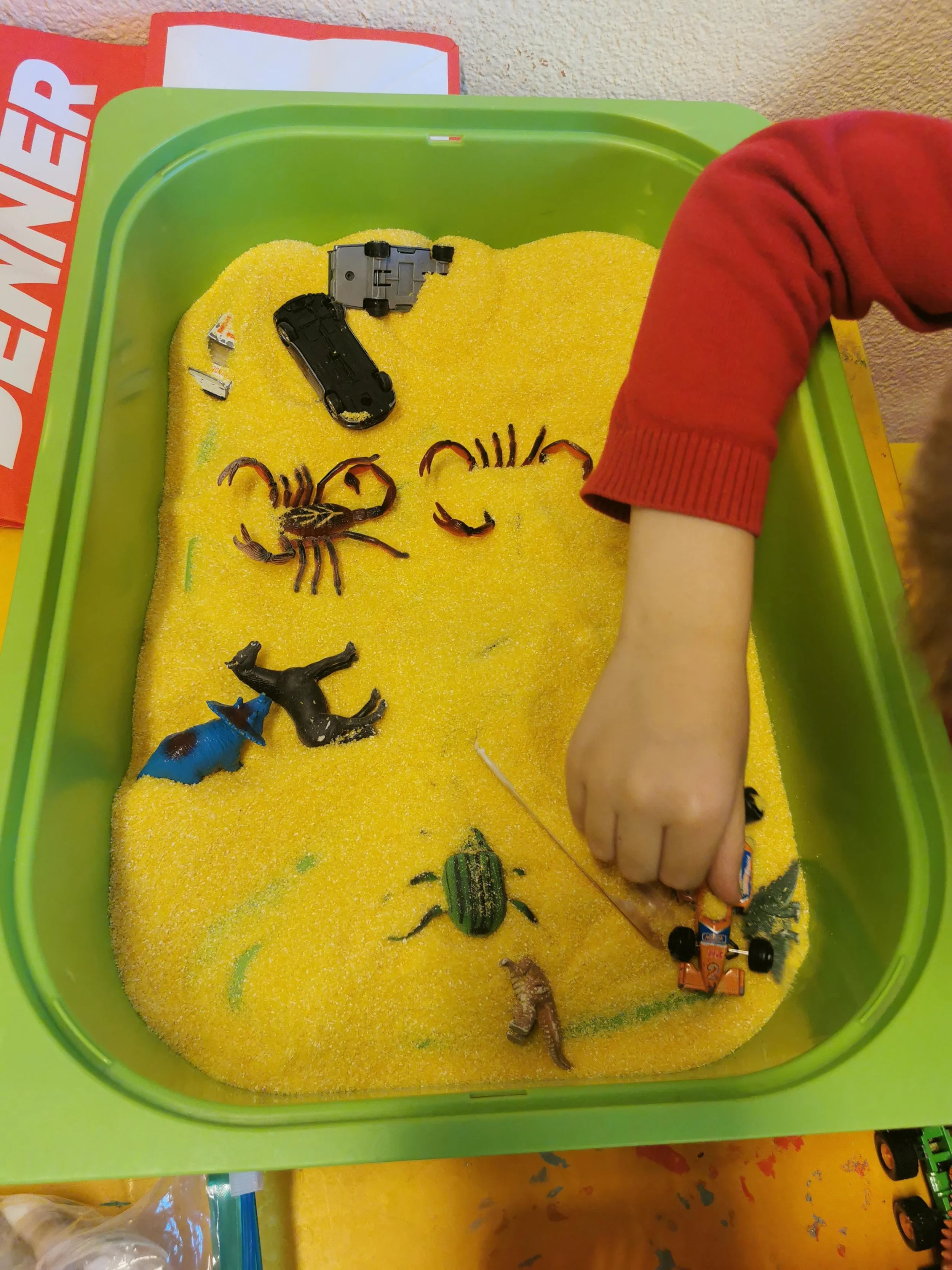 Child playing with toy insects and vehicles in a green sandbox filled with yellow sand.