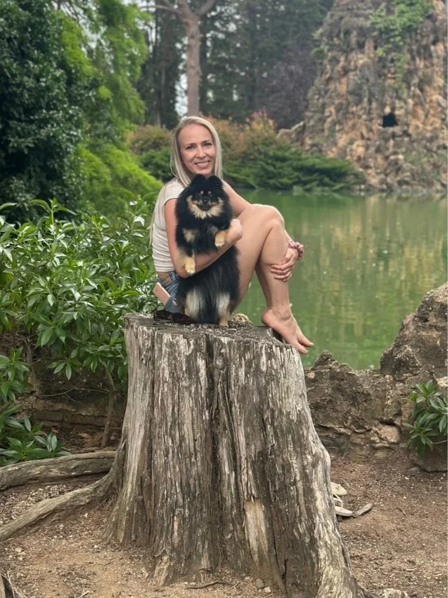 A woman sitting on a tree stump in a natural outdoor setting, holding a small black and tan fluffy dog. The background features green foliage, a calm body of water, and rocky cliffs.