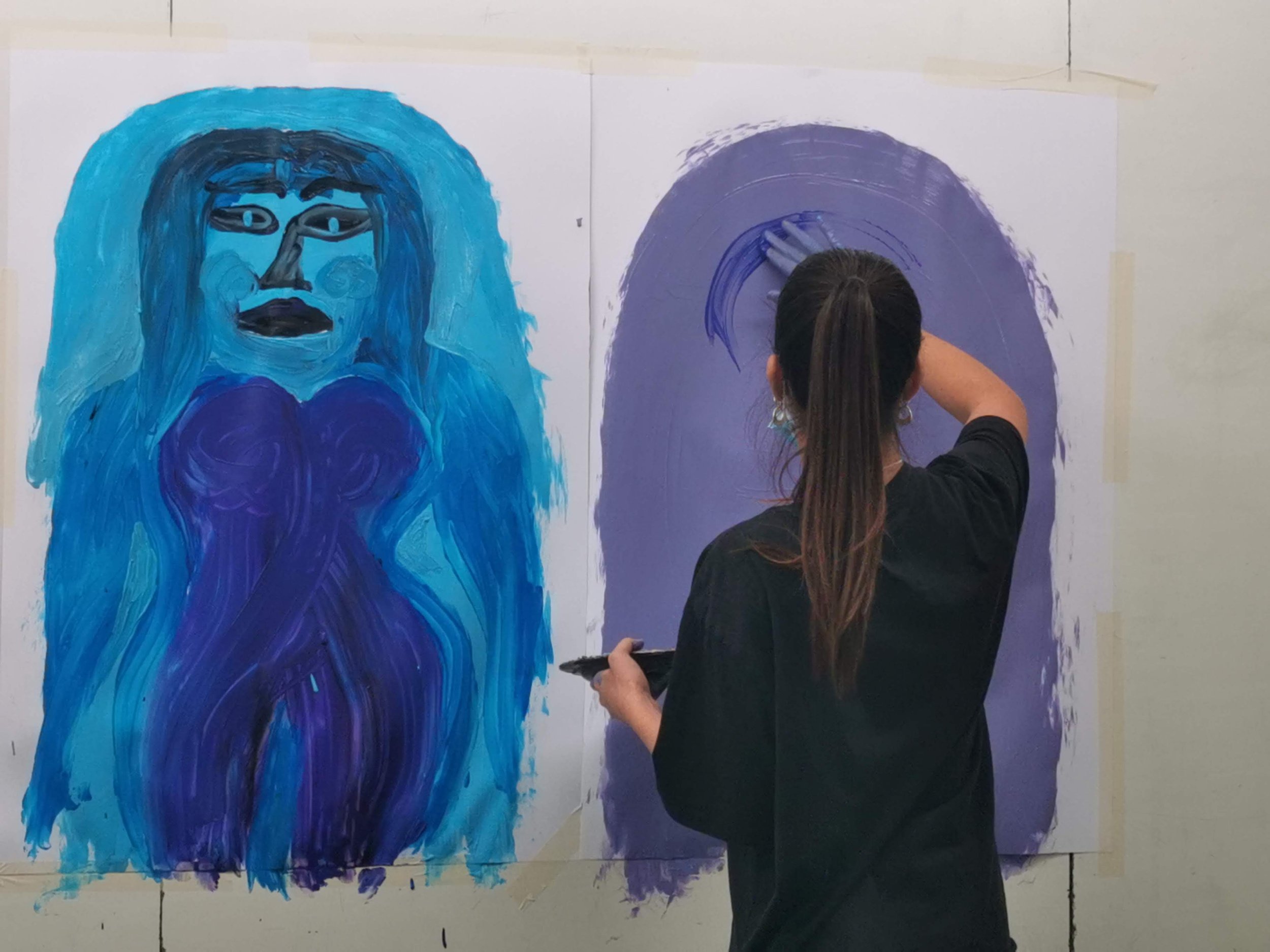 An artist with long brown hair tied in a ponytail, wearing a black shirt, painting on a large canvas. The artwork features abstract figures in shades of purple and blue, with the artist adding details to the purple background.