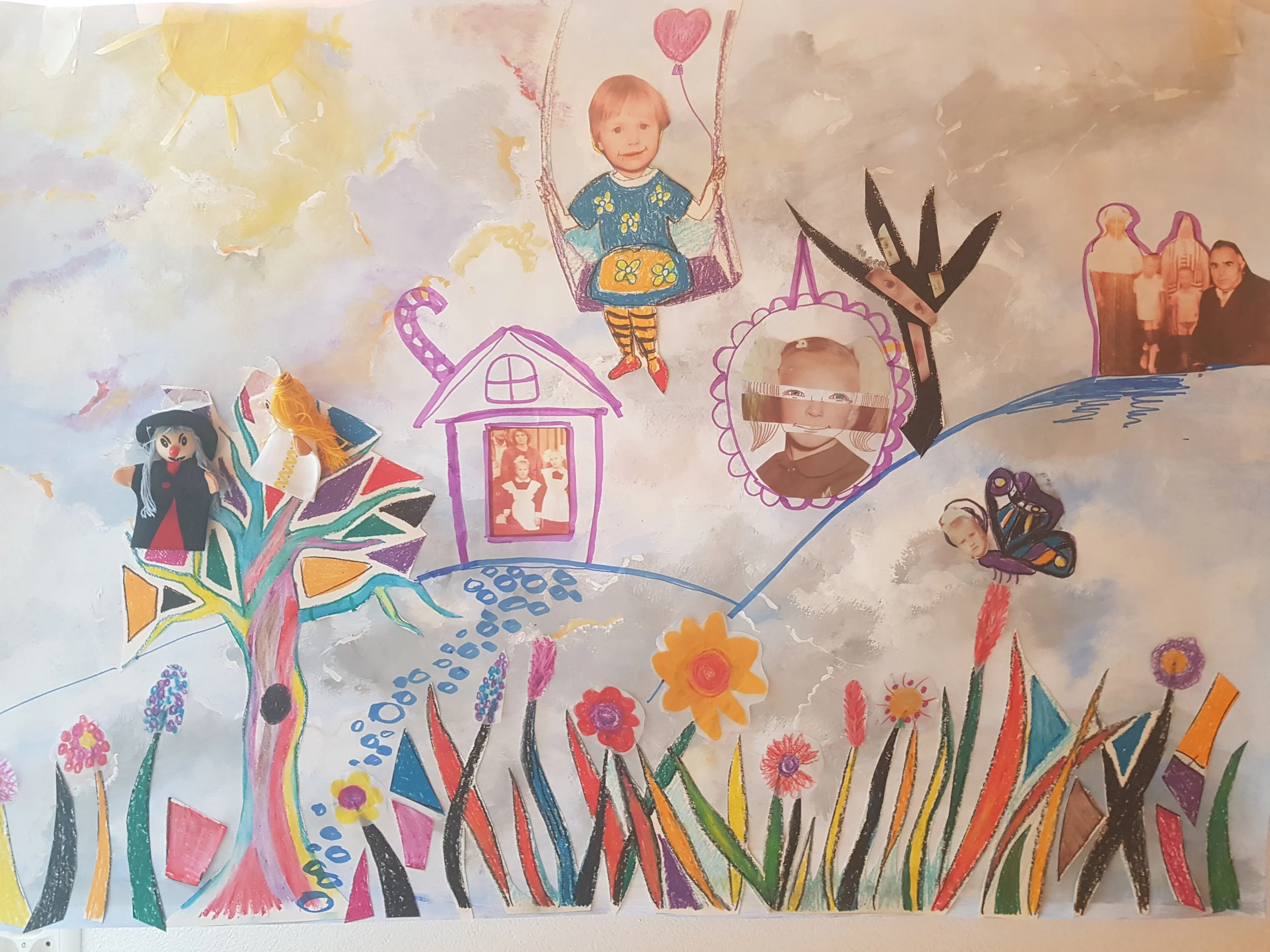 A colorful, hand-drawn collage on paper featuring a whimsical landscape with flowers, a tree, and a house, decorated with photographs of children and a family, along with drawings of a sun, butterfly, and various decorative elements.