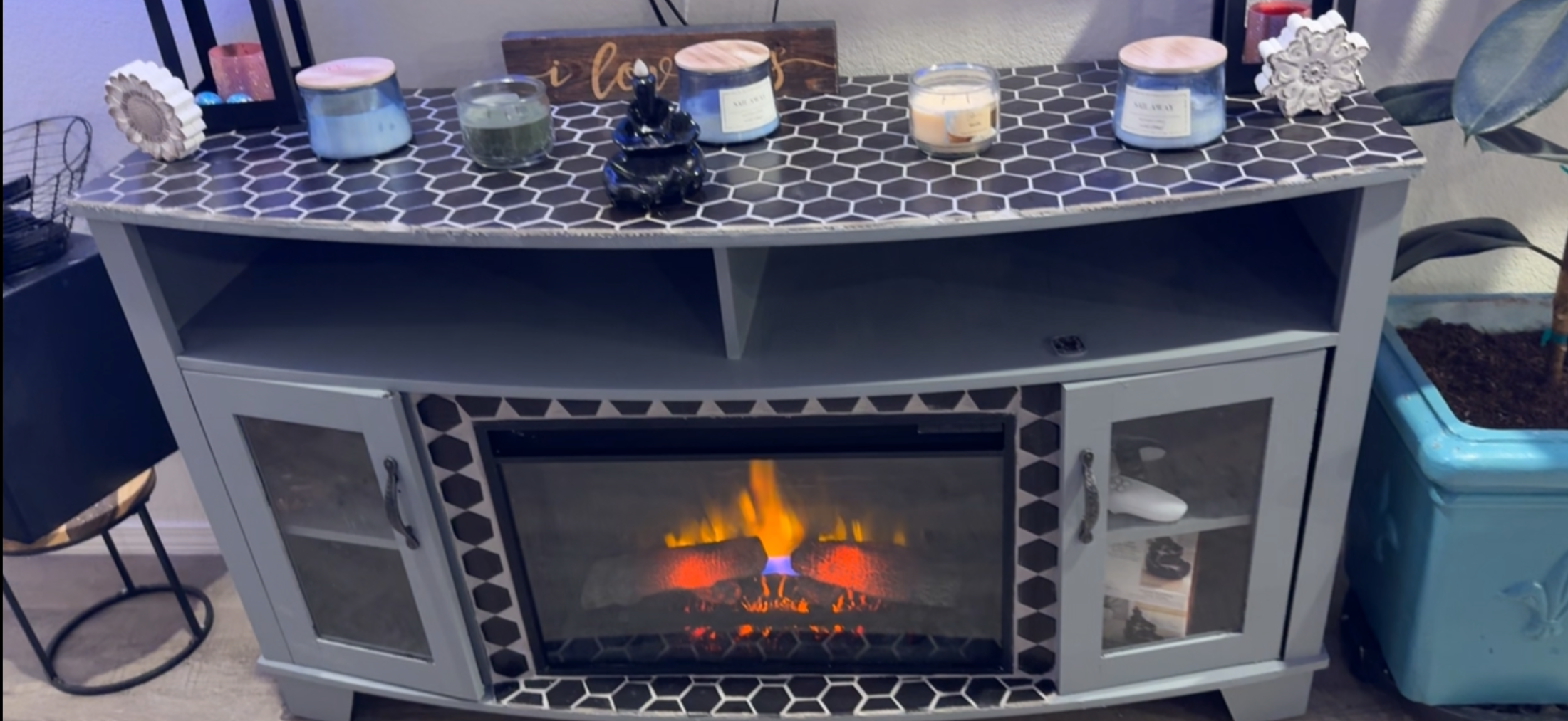 Gray entertainment console with hexagonal patterned black and white tiled top, an electric fireplace in the center, and glass doors on either side containing various items. Decor items including candles, a Buddha statue, and decorative objects are arranged on top.