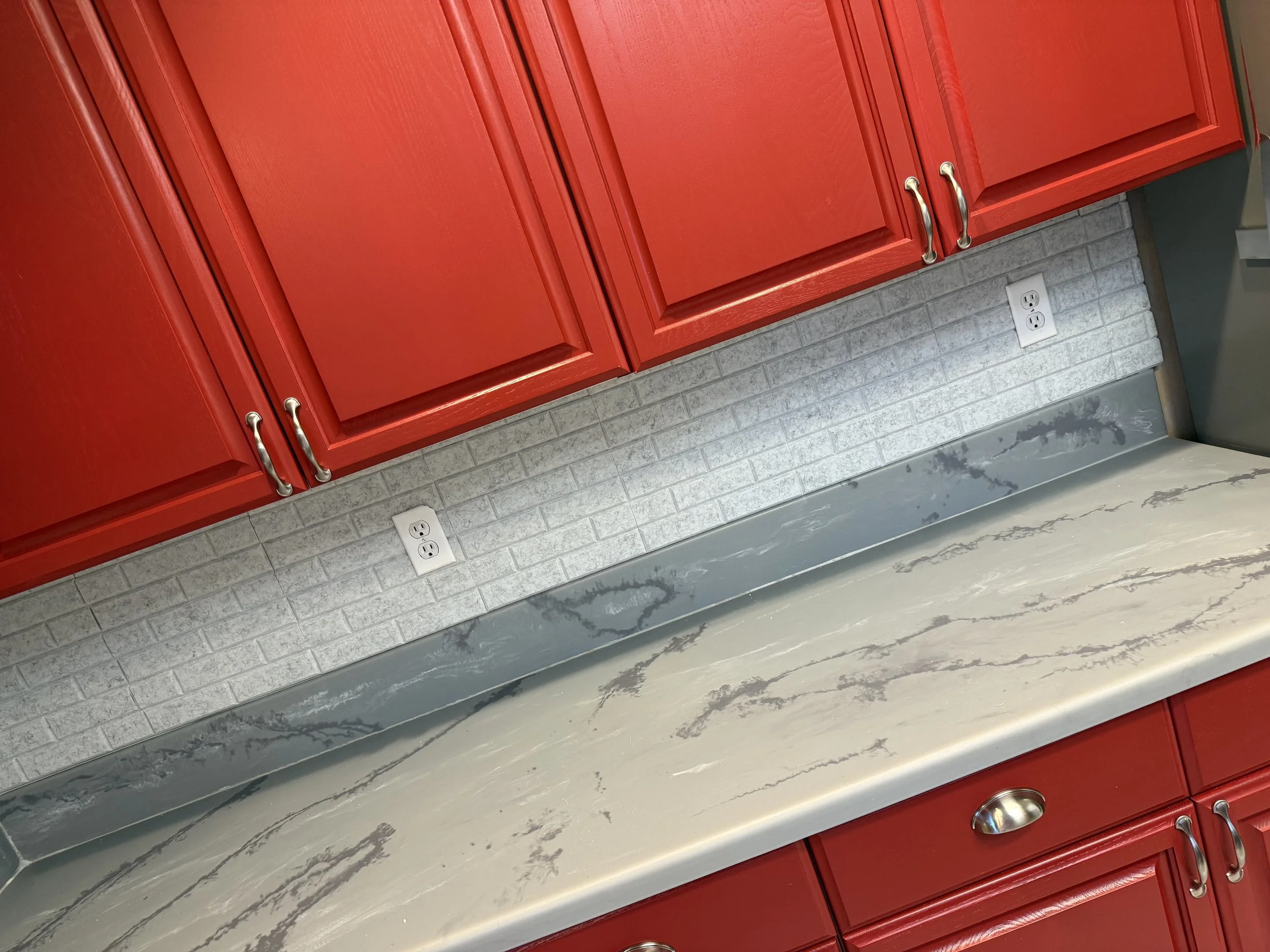 Red kitchen cabinets above a gray marbled countertop with a silver backsplash, white electrical outlets, and a gray wall.