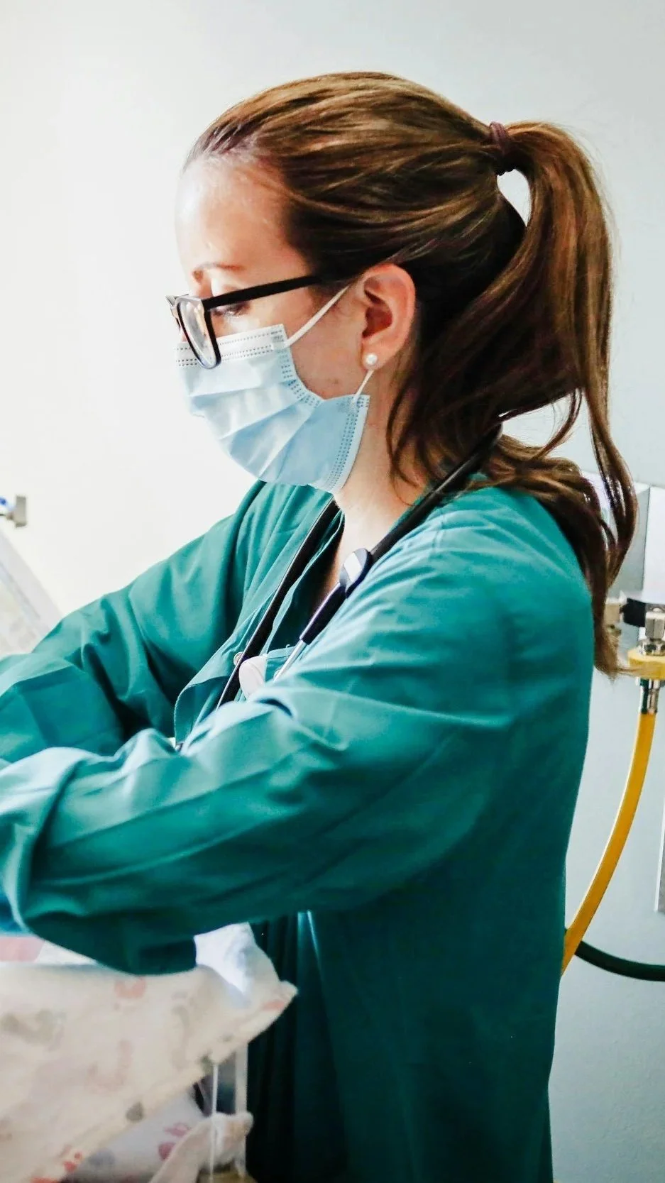 A female healthcare worker wearing a face mask, glasses, and green scrubs, working in a hospital.