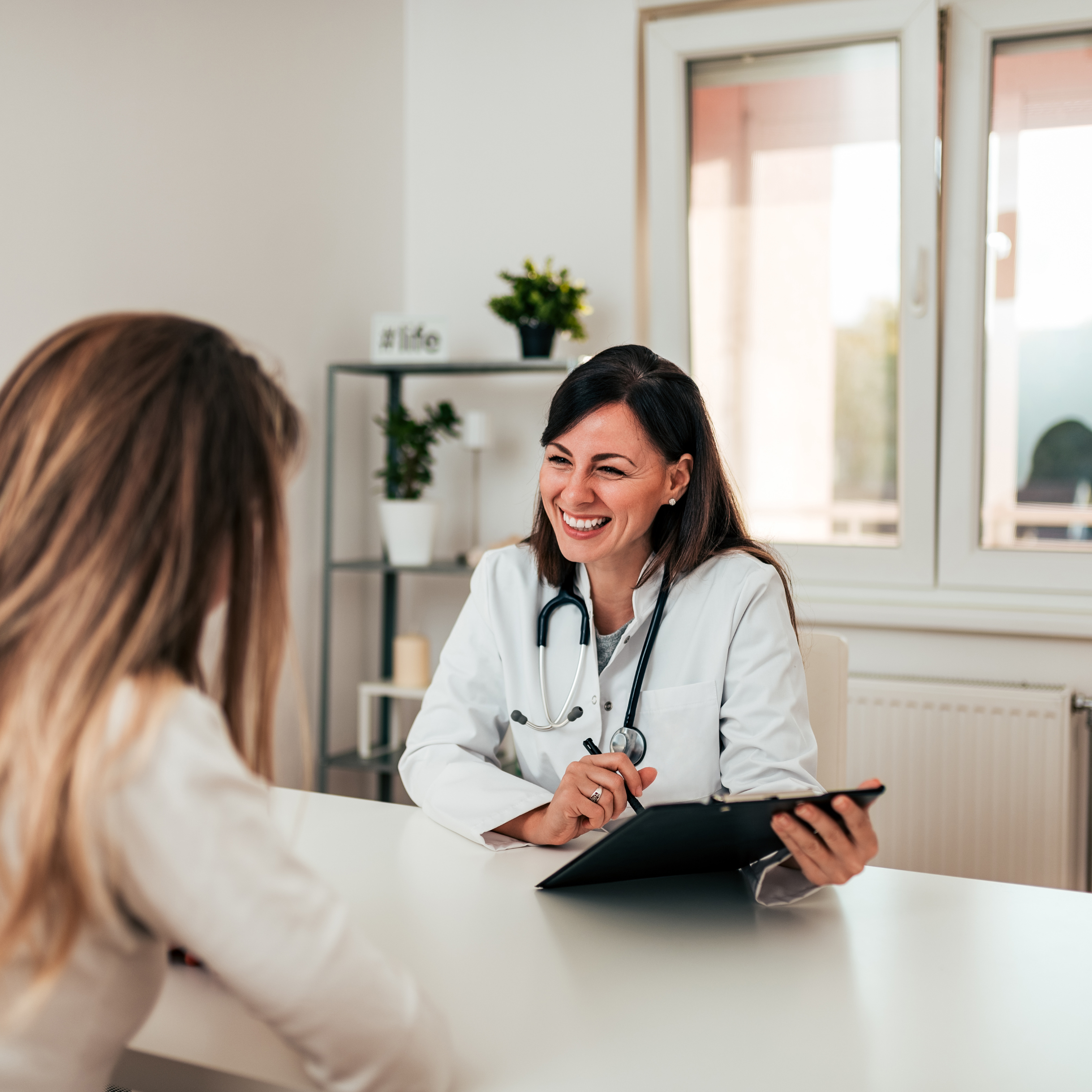 A female doctor in a white coat with a stethoscope around her neck is talking to a patient in a consultation room with white walls and a window. There are decorative plants and a small sign that says #life in the background.
