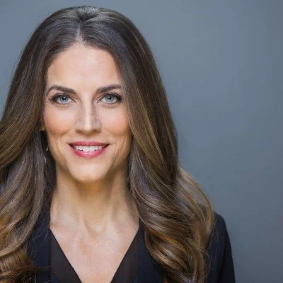 A smiling woman with long, wavy brown hair, wearing a black top against a plain gray background.