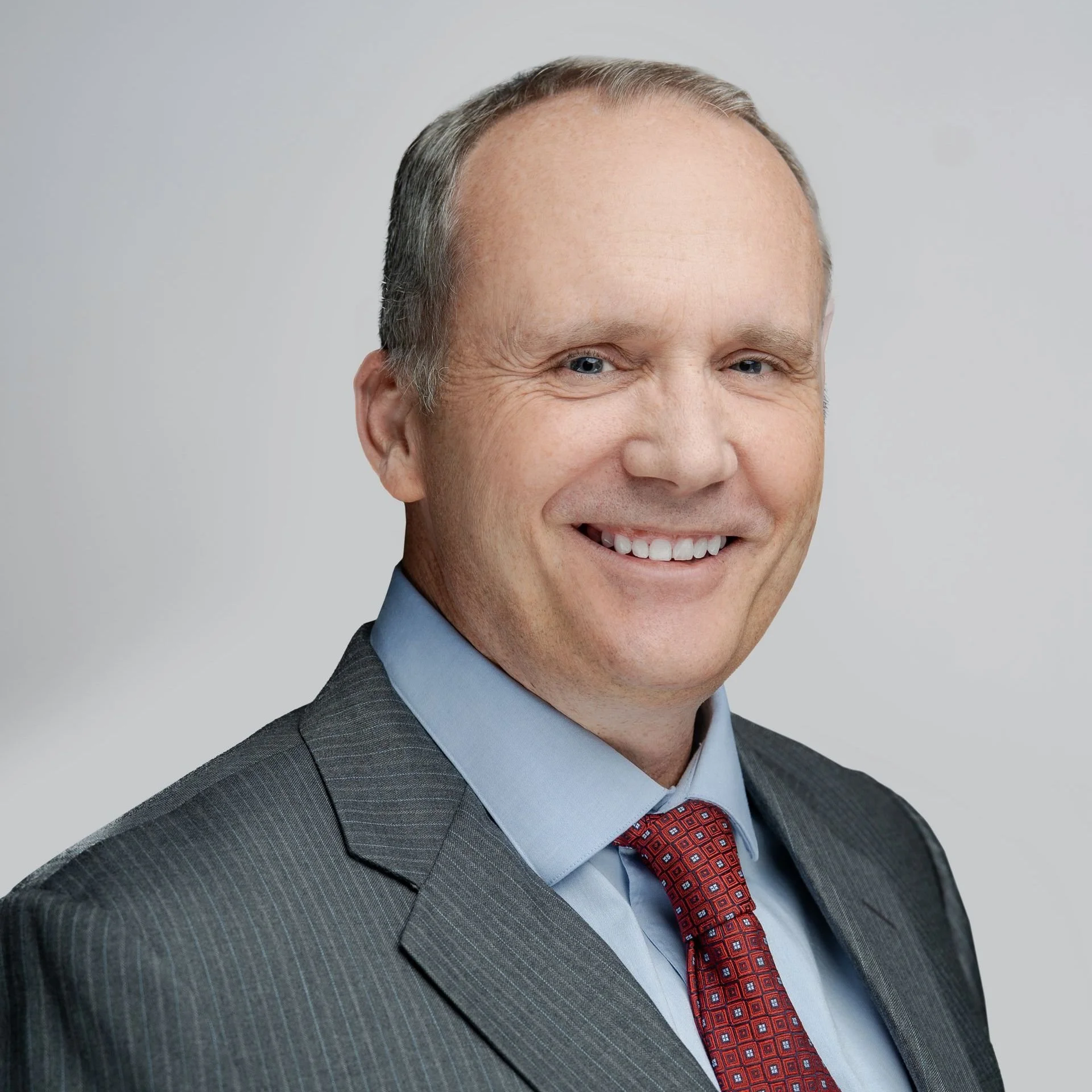 Smiling man dressed in a dark gray pinstripe suit, light blue shirt, and red patterned tie, against a plain light gray background.