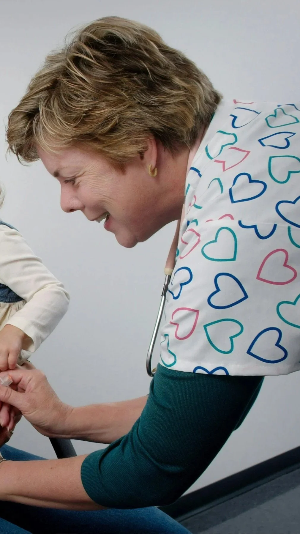 A female healthcare worker with short, light brown hair, wearing heart-patterned scrubs, smiling and interacting with a child in a medical setting.