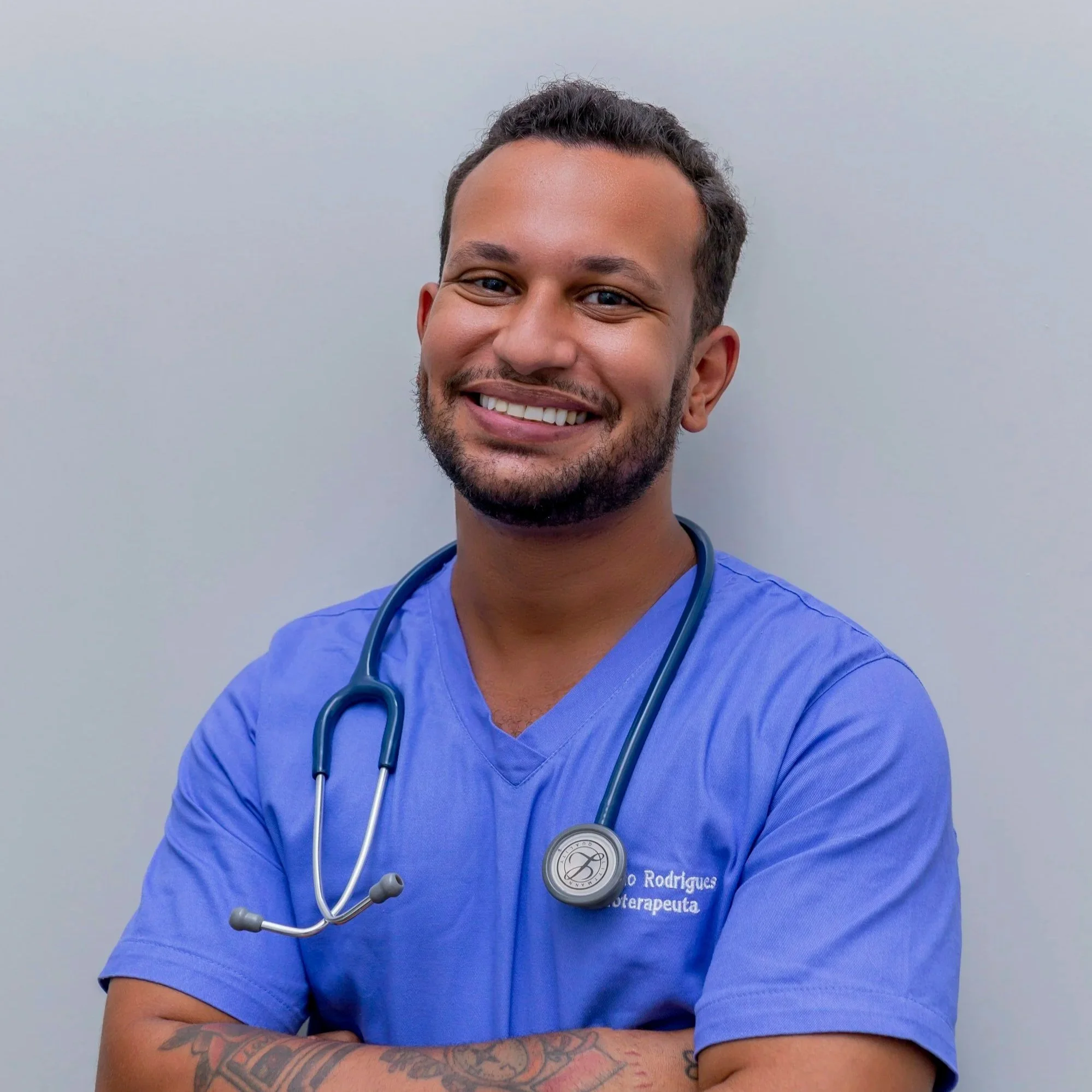Close-up portrait of a smiling male healthcare professional wearing blue scrubs and a stethoscope around his neck, standing against a plain light gray background.