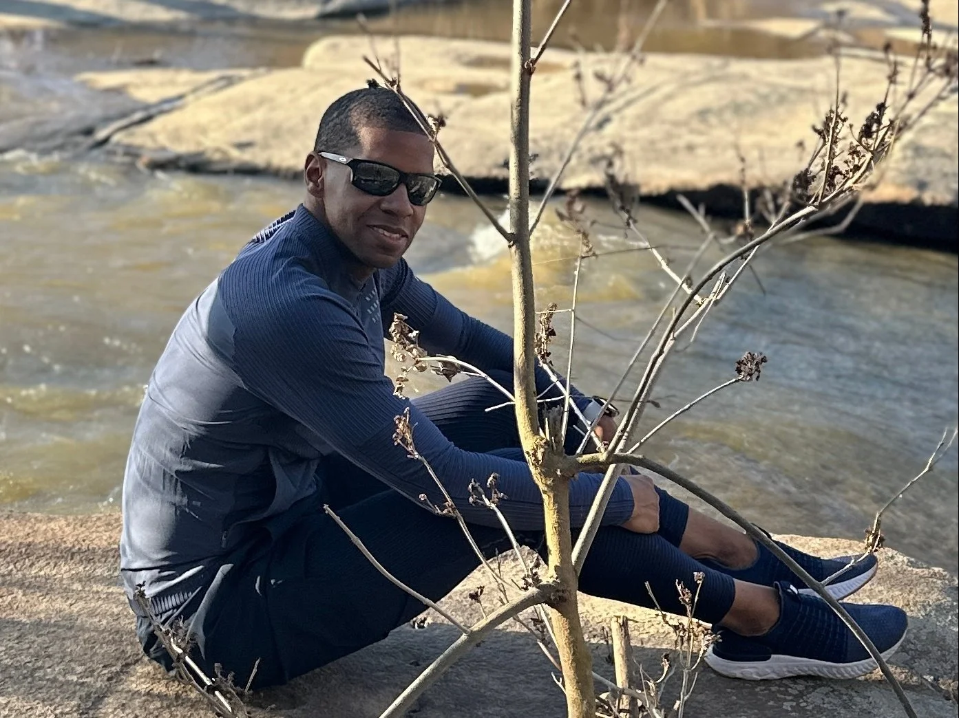 A man in athletic clothing wearing sunglasses sitting next to a river with a small tree and rocks in the background.