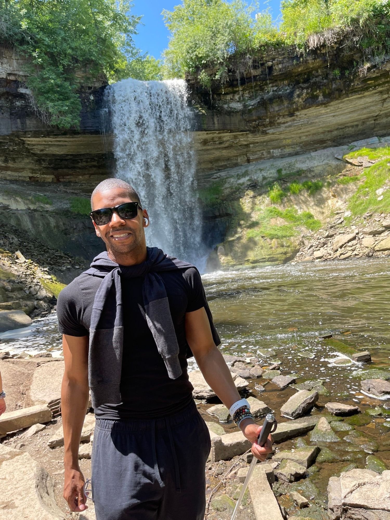 A man standing outdoors near a waterfall with green trees and rocks, holding a selfie stick, wearing sunglasses, a black shirt, and a gray sweatshirt tied around his neck.