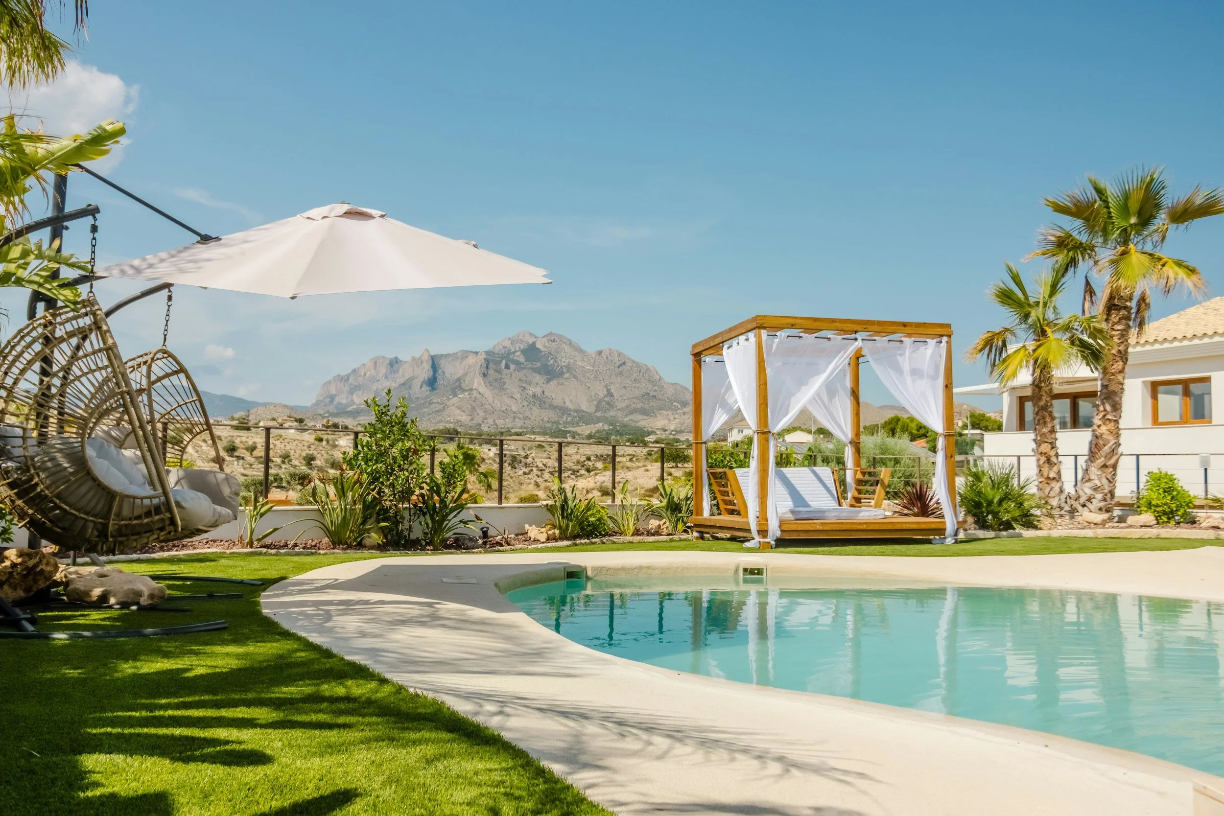 Luxury backyard with swimming pool, sitting area with hanging chairs, cabana, umbrellas, palm trees, and mountains in the background under a clear blue sky.
