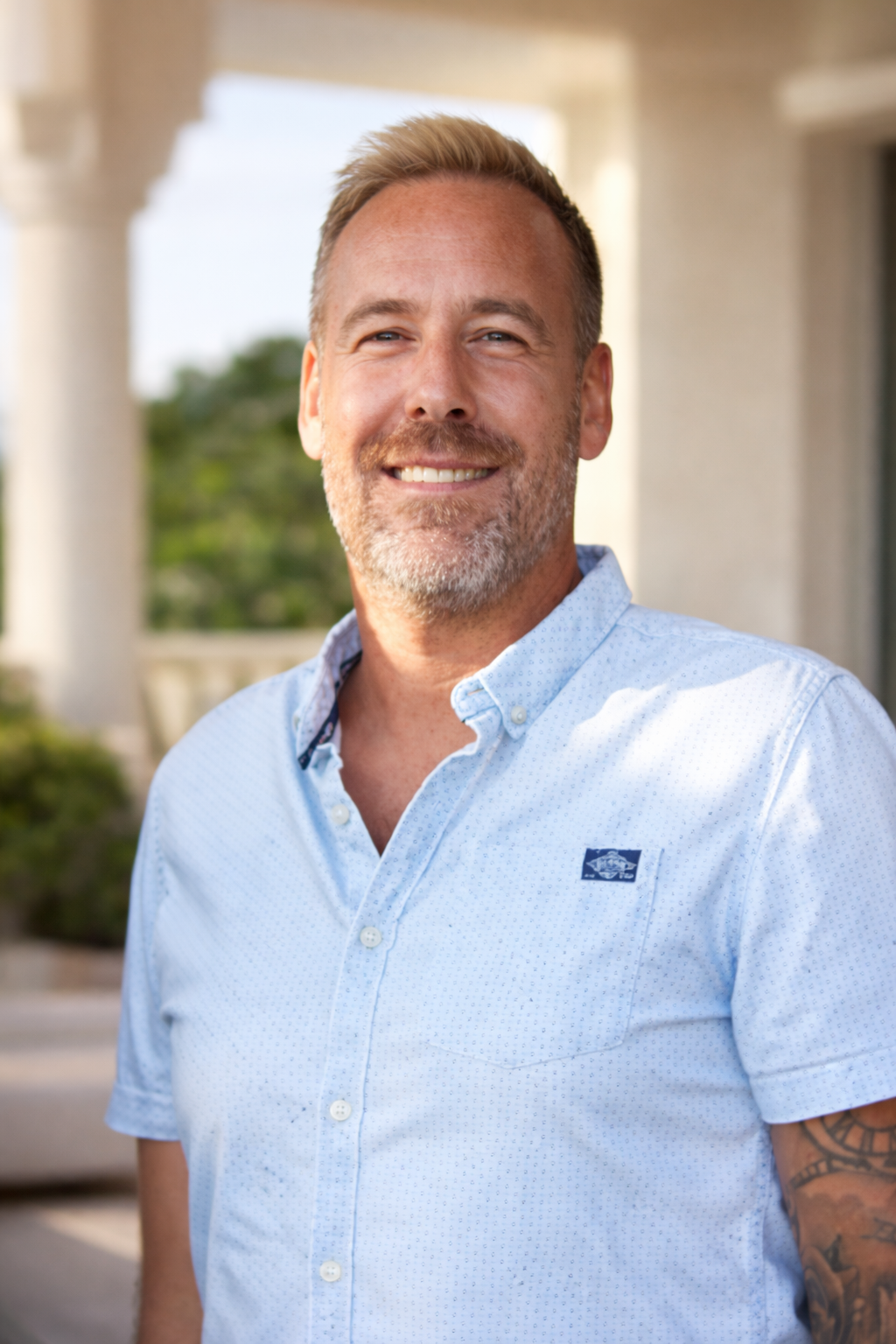 A smiling man with light skin, short blond hair, and a beard, wearing a light blue button-up shirt, standing outdoors in front of a white building with columns and greenery.