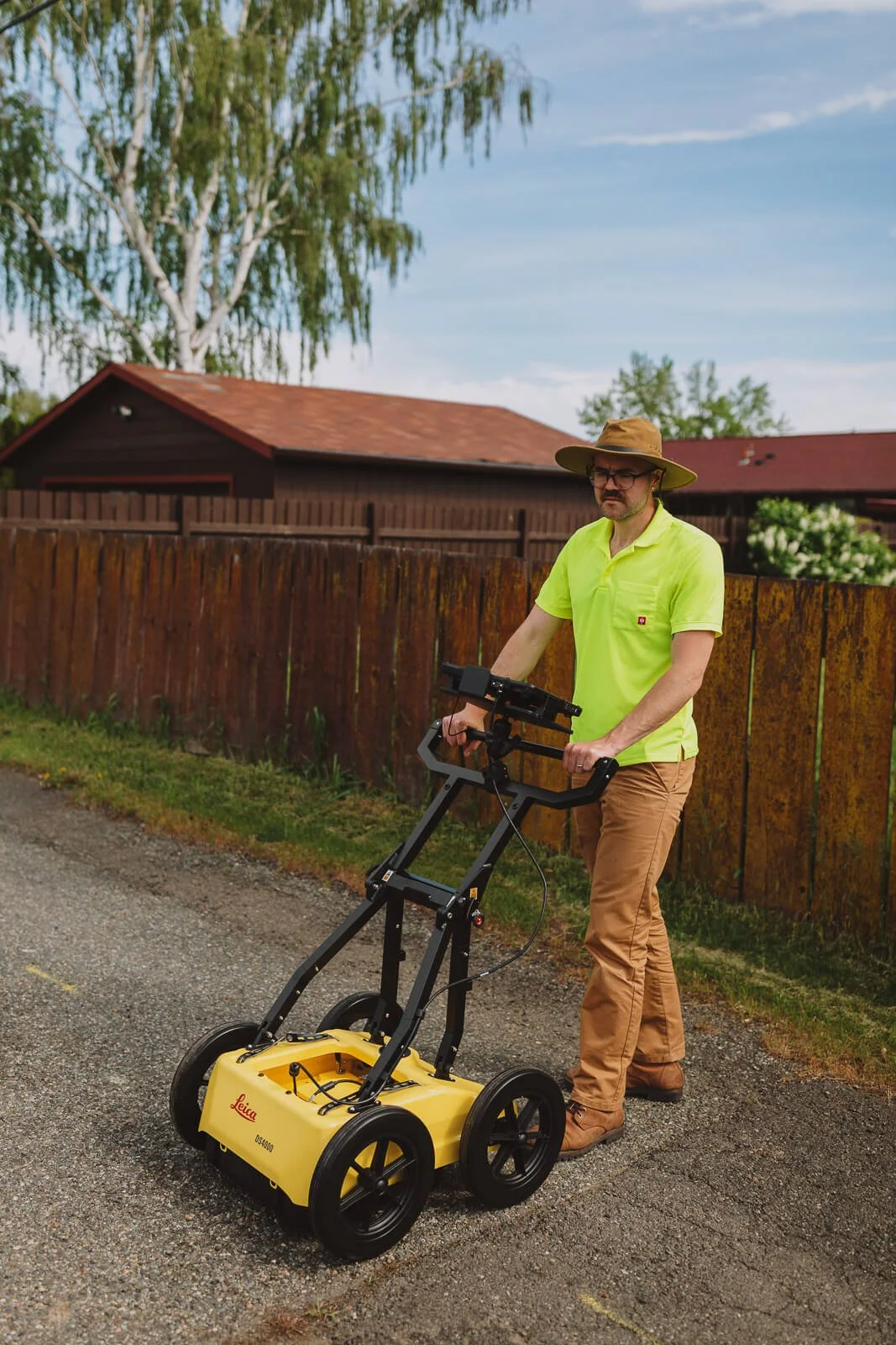 A man wearing a wide-brimmed hat, glasses, a neon yellow polo shirt, and tan pants is standing on a paved road, operating a yellow Leica ground-penetrating radar device. There is a wooden fence, trees, a house with a red roof, and a partly cloudy sky in the background.