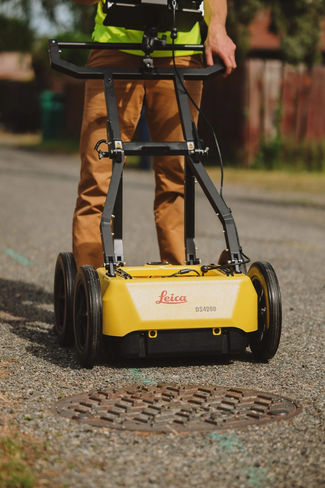 A person operating a yellow Leica 3D Scanner on wheels outdoors on a gravel surface.