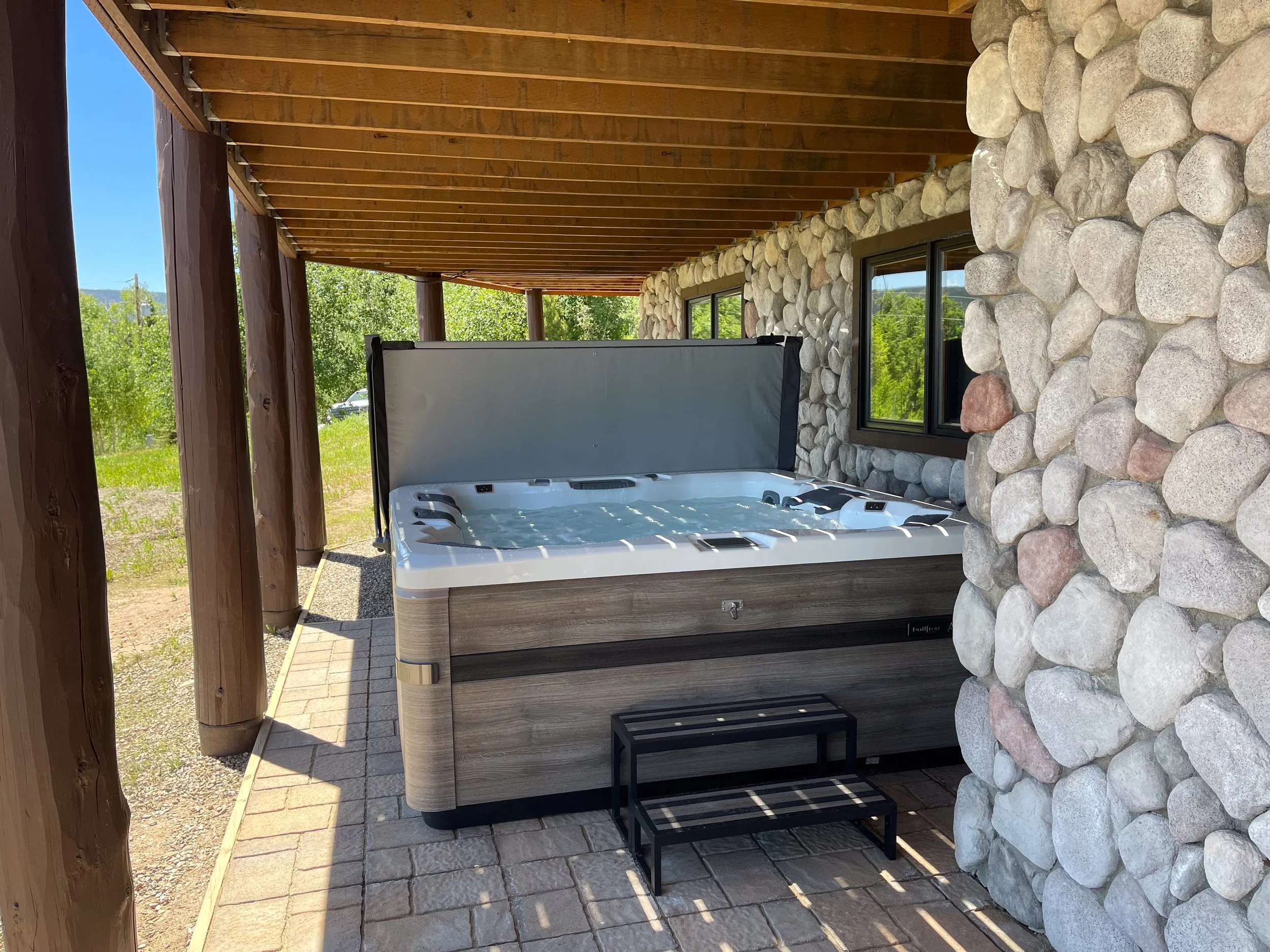 Outdoor hot tub on a patio with stone wall, window, wooden roof, and greenery in the background.