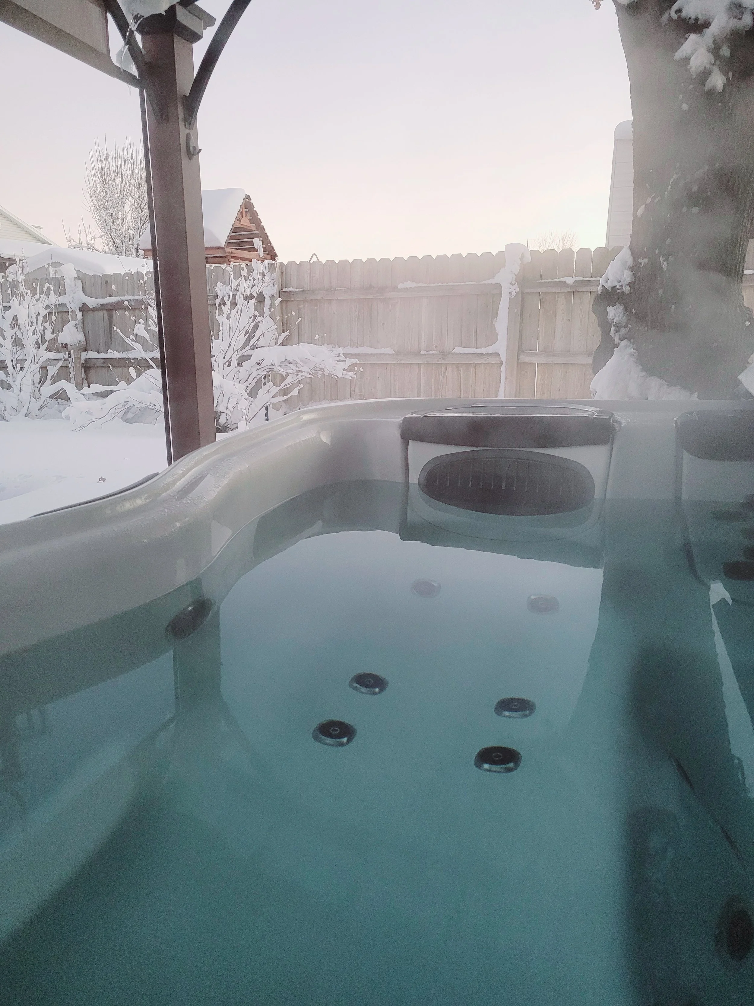 An outdoor hot tub filled with water, with snow-covered trees and a wooden fence in the background during winter.