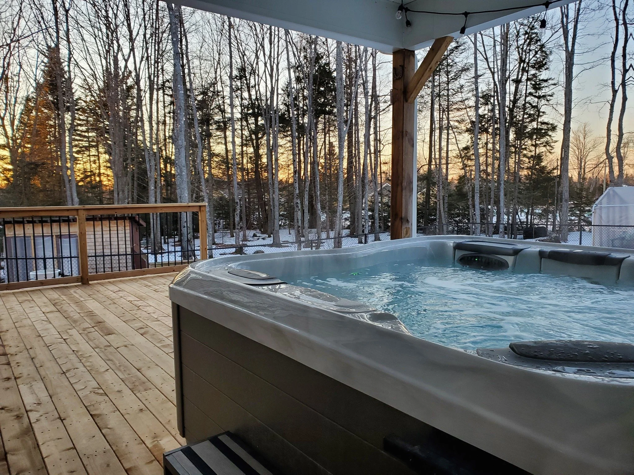 A hot tub on a wooden deck overlooking a snowy wooded yard during sunset.